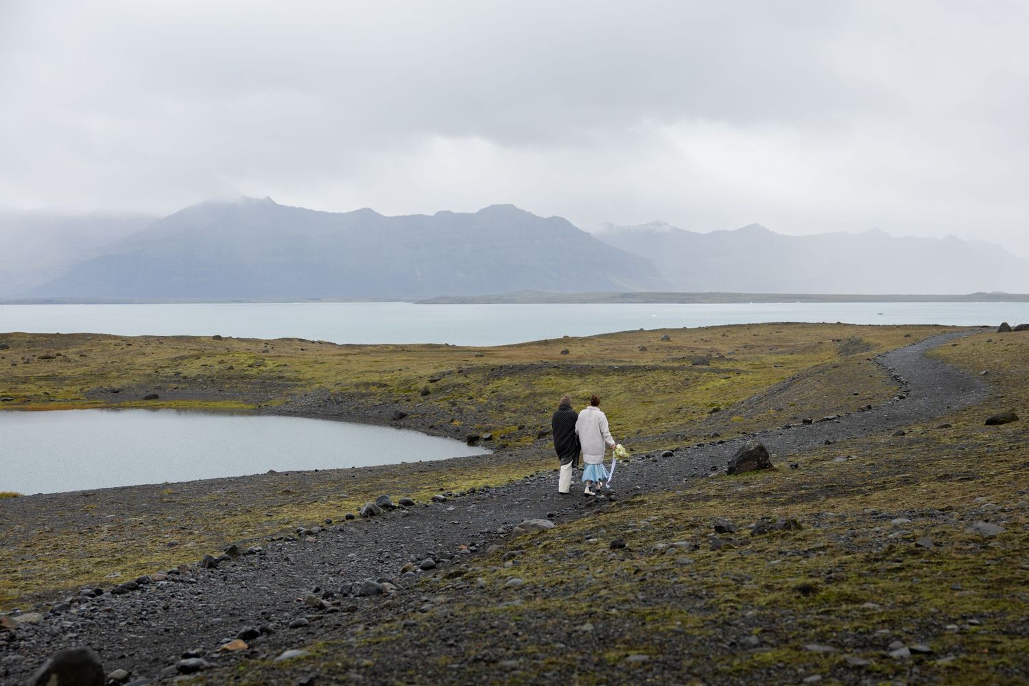 Two brides walking on a rocky path to Jokulsarlon glacier lagoon in Iceland with misty mountains in the background by Taylor Zimmerman Photo