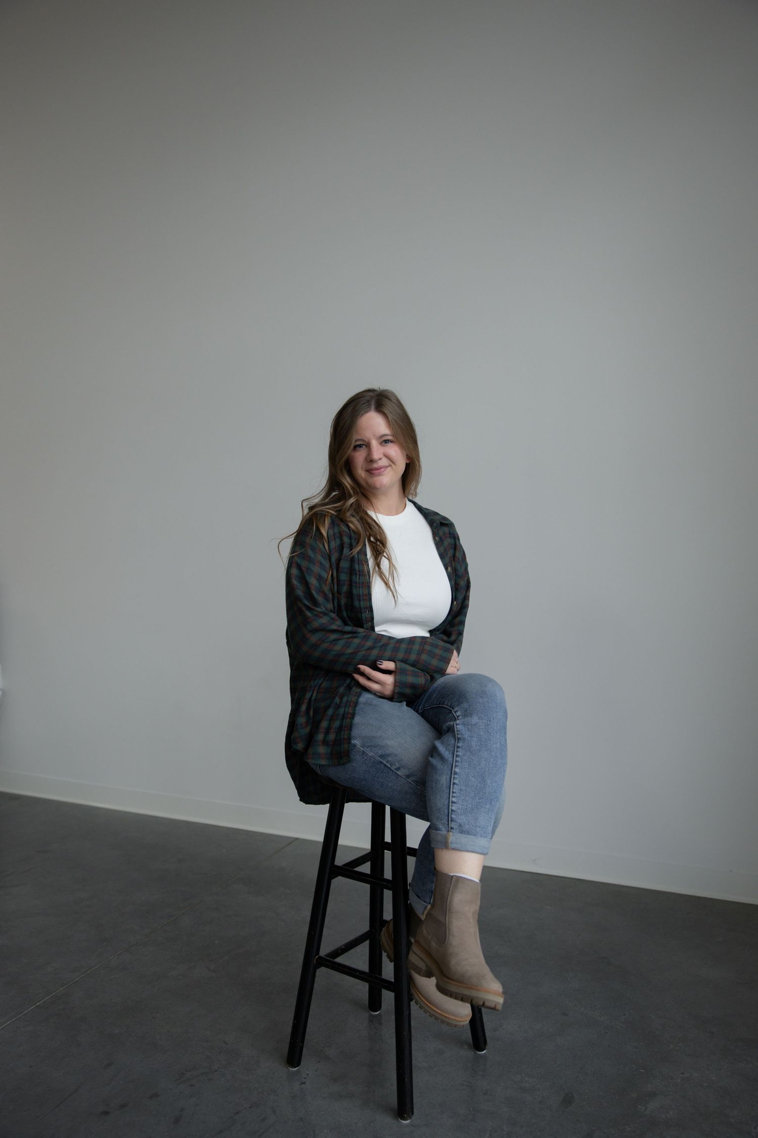Taylor Zimmerman in plaid shirt, white tee, jeans, and boots sitting on a black stool against a gray wall.