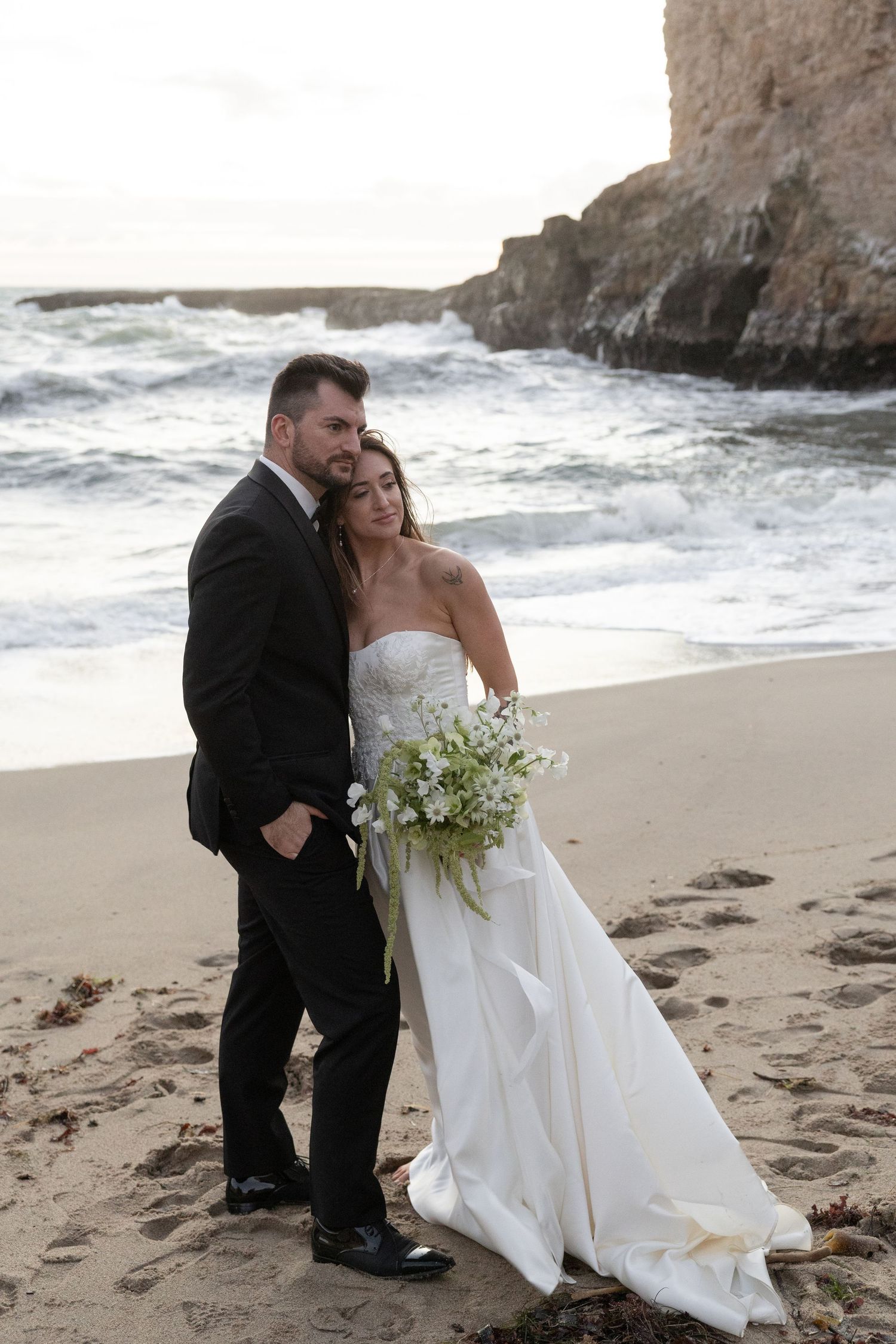 Bride and groom pose for a portrait on a sandy beach with ocean waves and rocky cliffs, bride holding white floral bouquet.