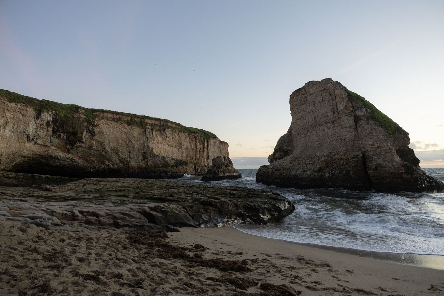 Rocky coastal beach at Shark Fin Cove with dramatic sea cliffs and large shark fin formation at dusk, with gentle waves on sandy shore.