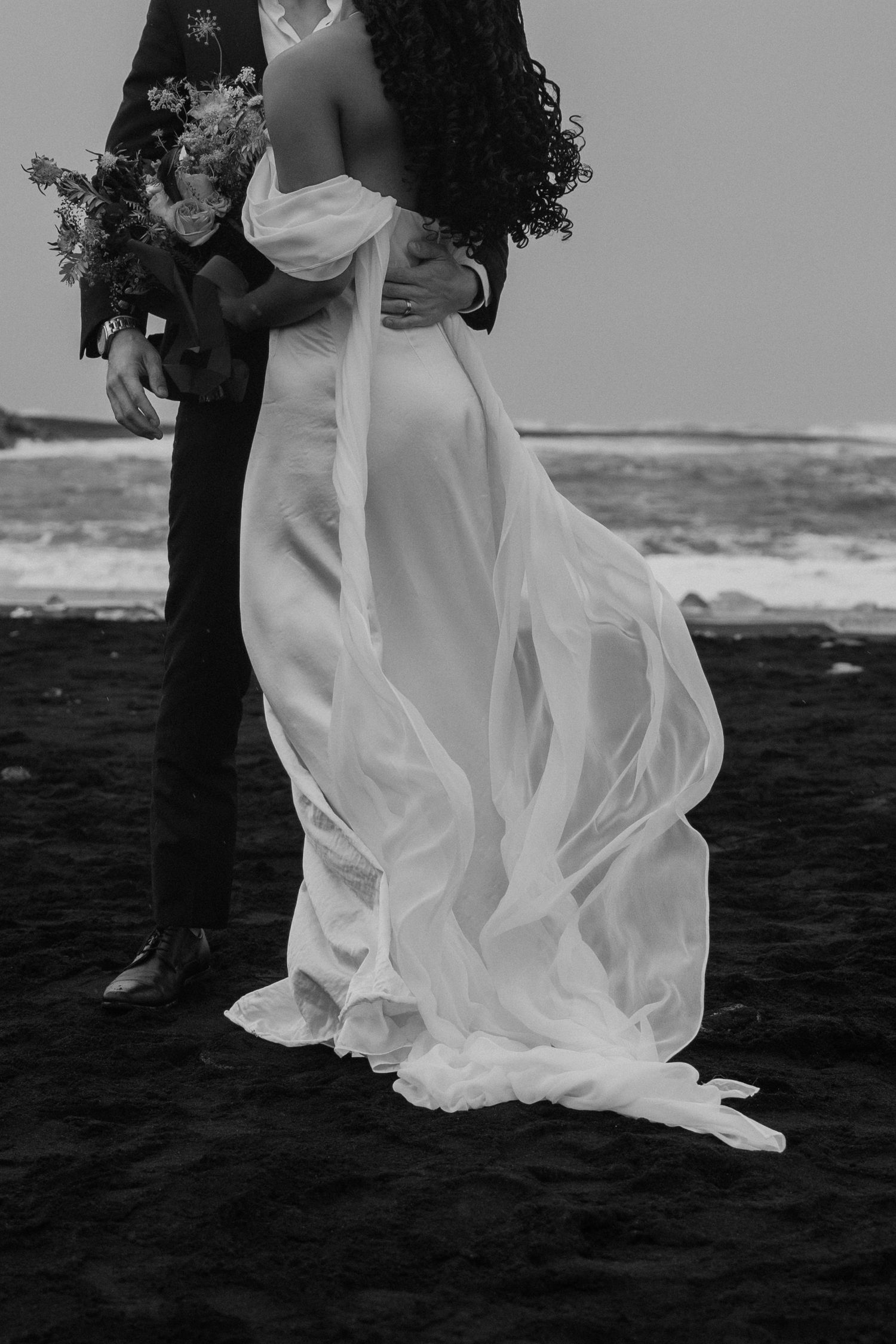 The bride's dress flowing in the Iceland wind while hugging her husband at Breidamerkursandur Beach in black and white by Taylor Zimmerman