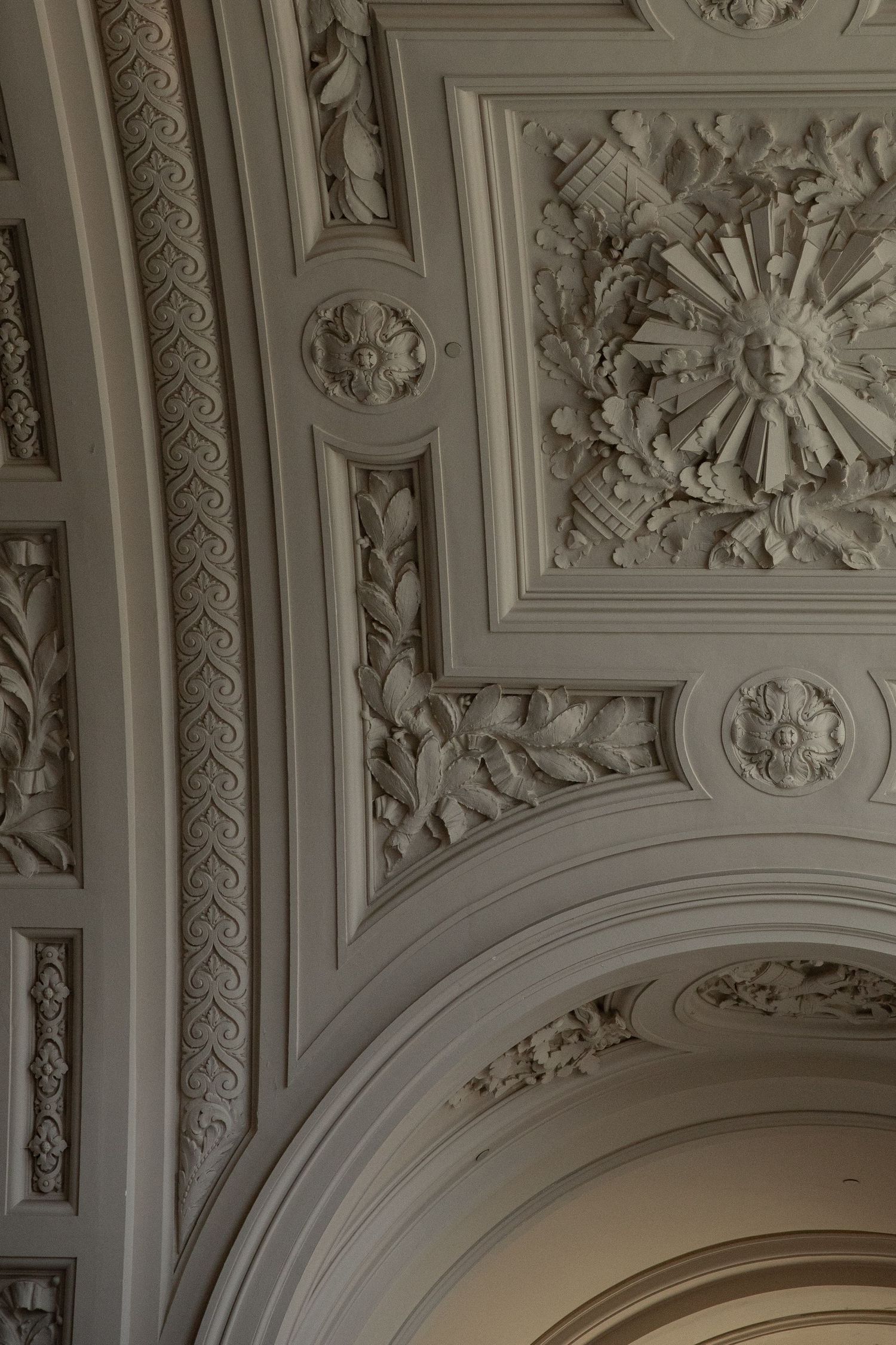 Ornate white plaster ceiling with intricate floral carvings, sun motif relief sculpture, and elegant arched architectural details.