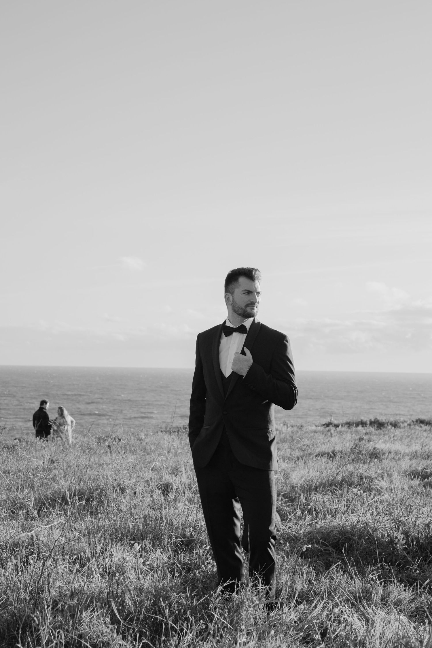 Photo of a groom in a tuxedo standing in a coastal field, adjusting his bow tie in black and white.