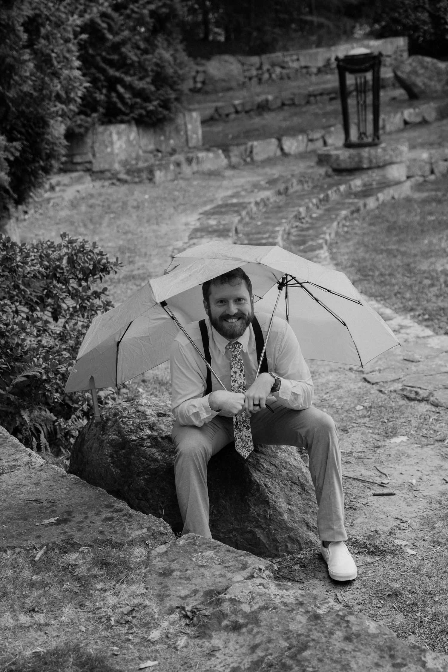 A smiling bearded man in suit sitting on a rock holding two umbrellas above his head in black and white by Taylor Zimmerman Photo