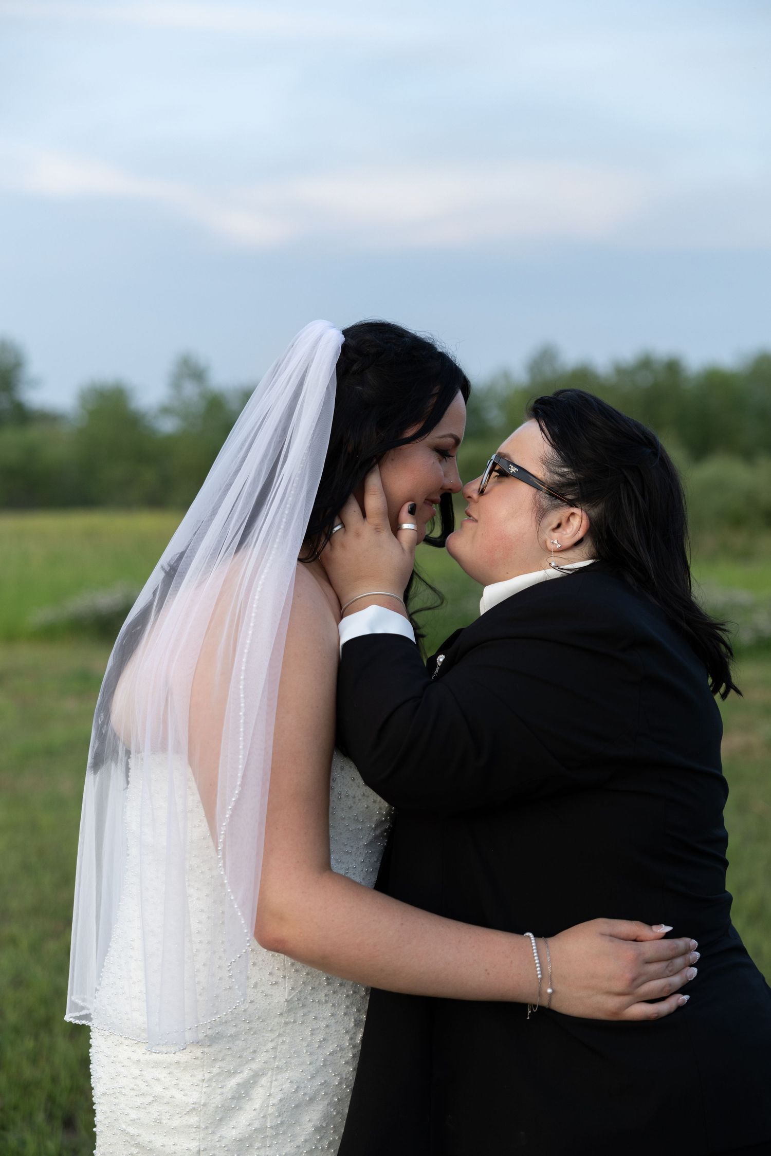Two brides lean in for a kiss, one in a white gown and veil, the other in a black suit, in an open field on their wedding day
