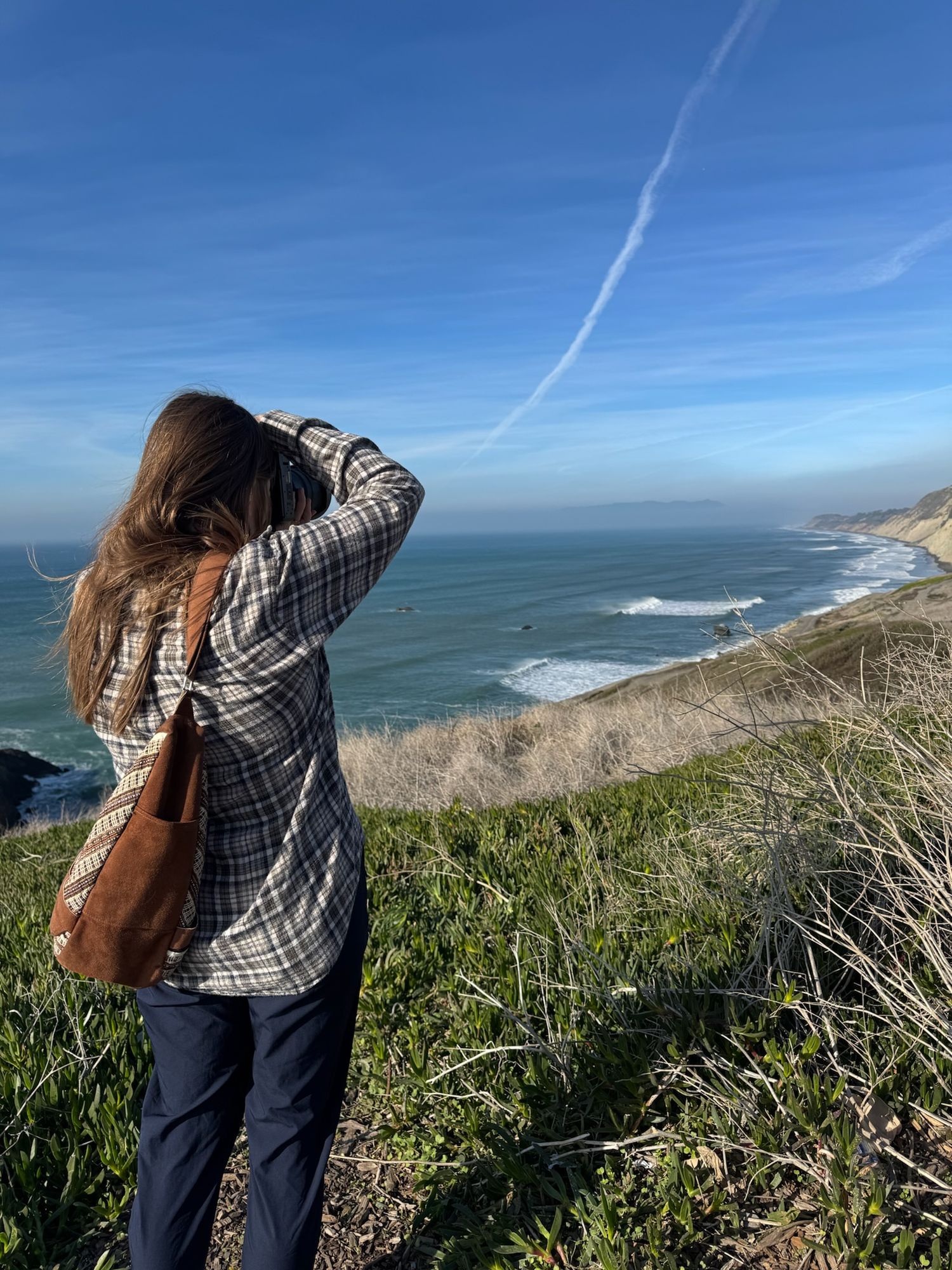 Woman in plaid shirt photographing scenic coastal cliffs and ocean waves under a clear blue sky