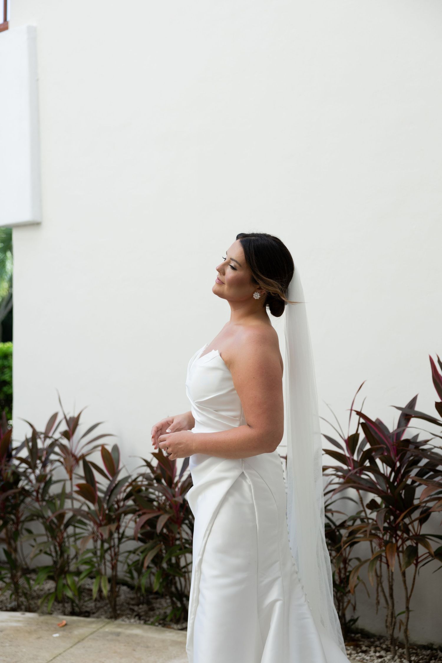 Bride in strapless white gown and veil poses gracefully against white wall surrounded by tropical foliage by Taylor Zimmerman Photo