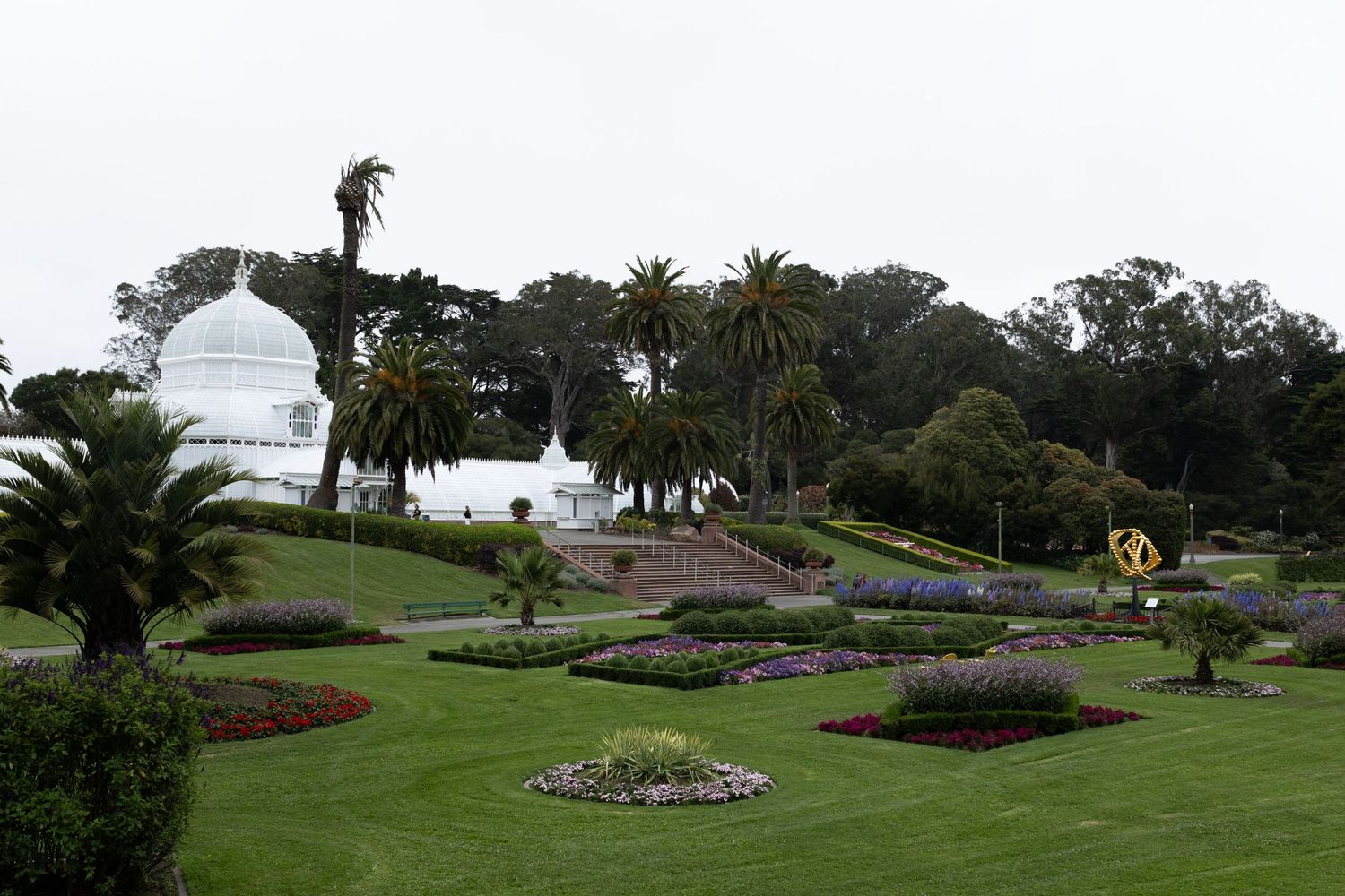 Lush garden landscape with colorful flower beds and palm trees near a white domed conservatory in Golden Gate Park, San Francisco.