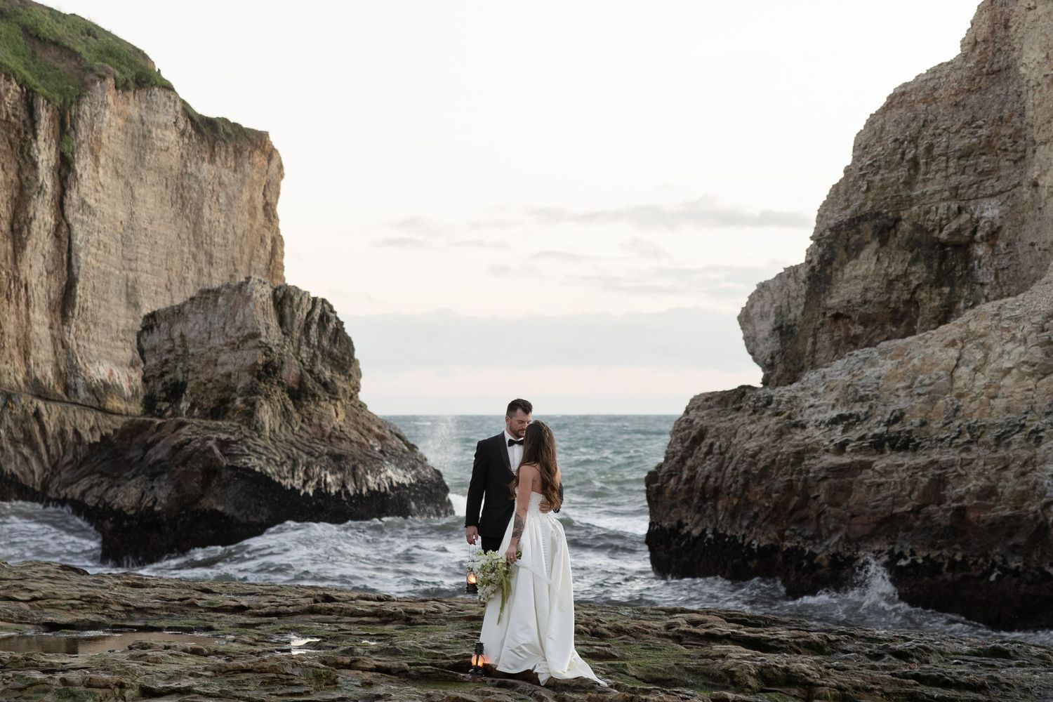 Bride and groom kissing on rocky beach with dramatic ocean cliffs at sunset during coastal wedding ceremony.