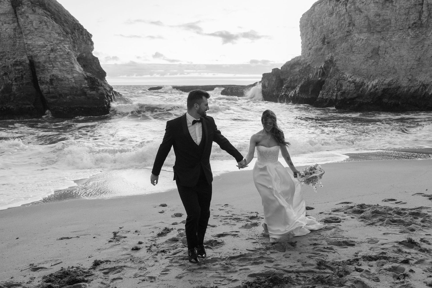 Photo of a bride and groom holding hands walking on a rocky beach with ocean waves at Shark Fin Cove in black and white.