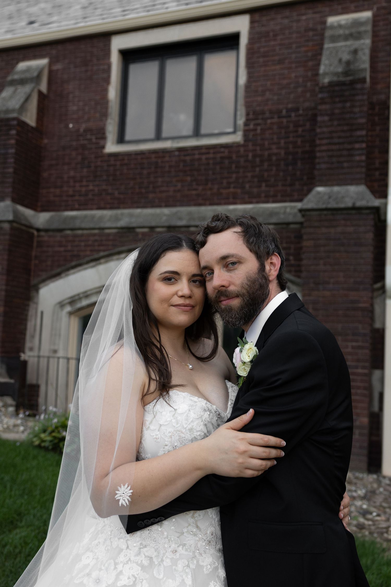 Bride and groom posing in front of a brick building, bride wearing lace gown and veil, groom in black suit by Taylor Zimmerman Photo