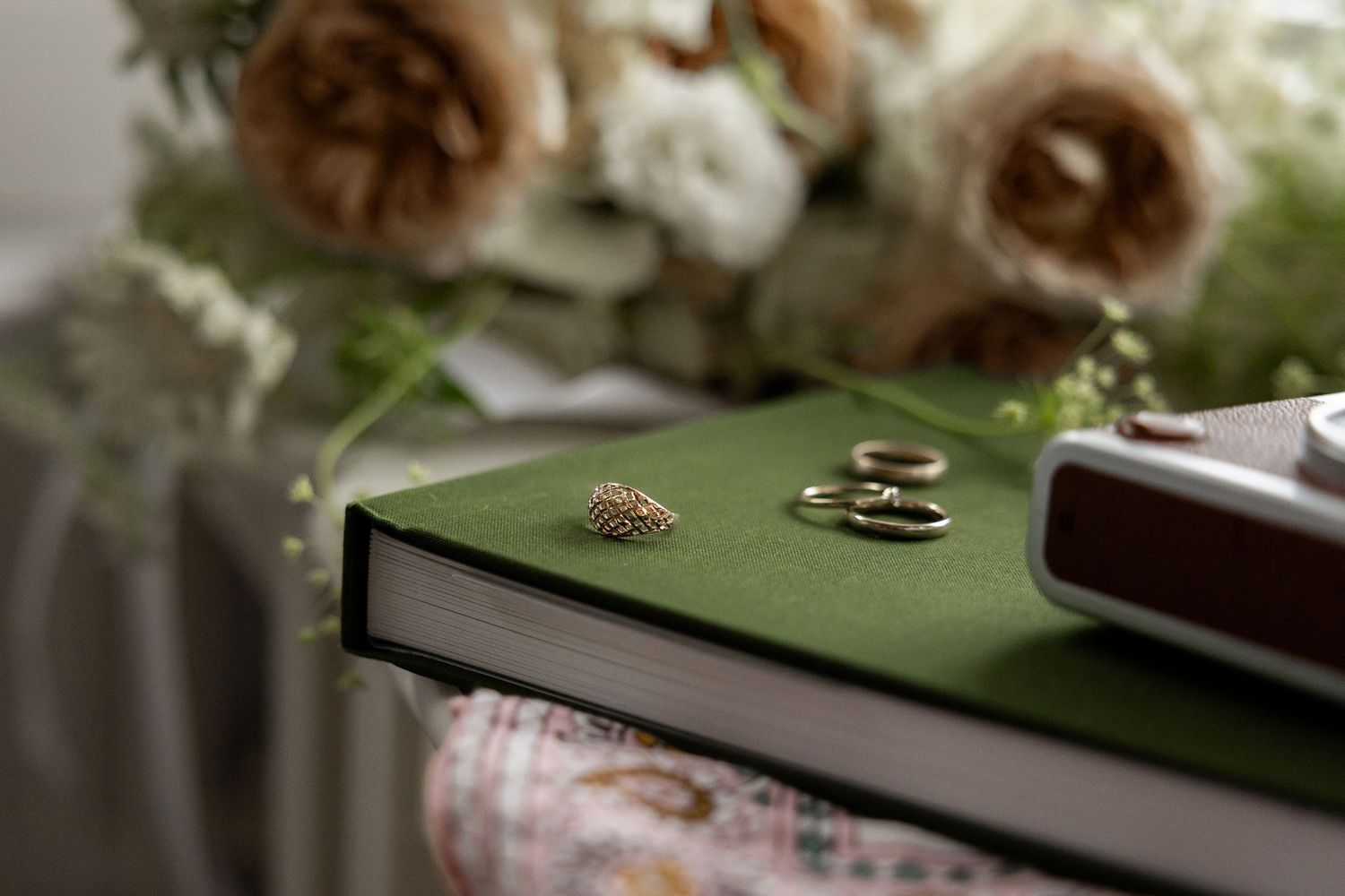Wedding rings and an heirloom ring rest on a green book beside a bridal bouquet of blush and white flowers by Taylor Zimmerman Photo