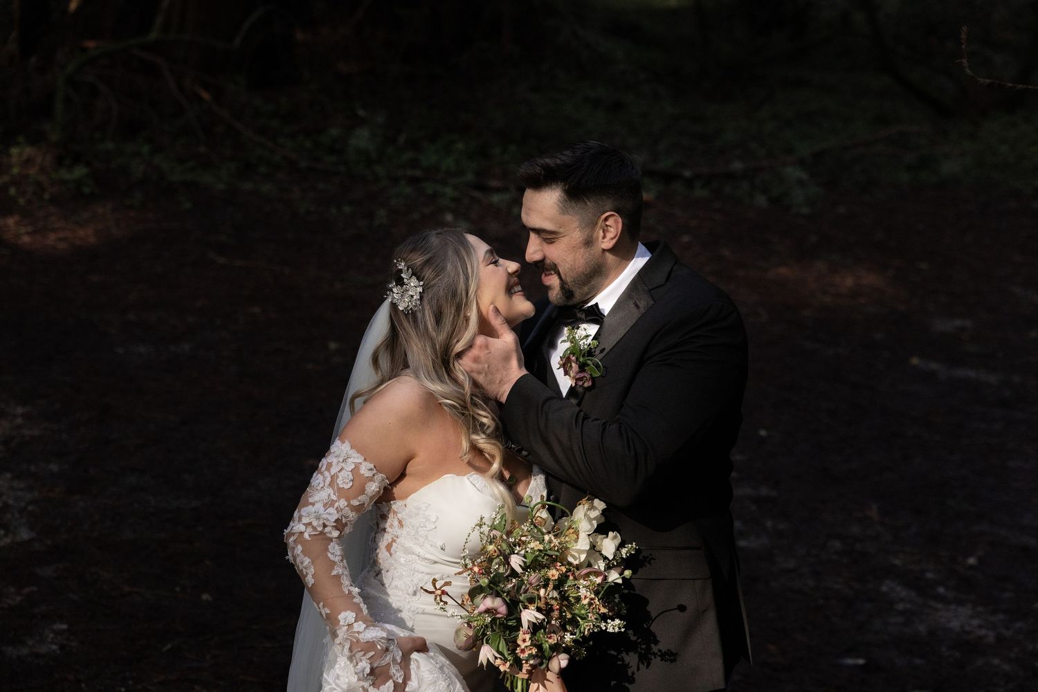 A couple share a romantic moment in a dark forest, with the bride holding a lush bouquet by Taylor Zimmerman Photo