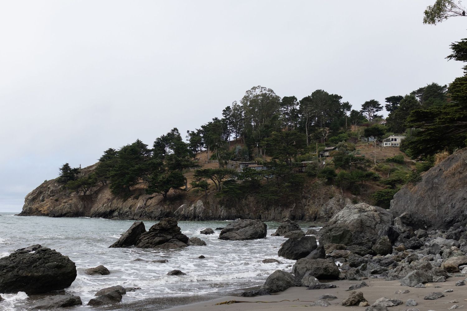 Rocky beach with dark sand, large boulders, and a tree-covered coastal cliff under an overcast sky.