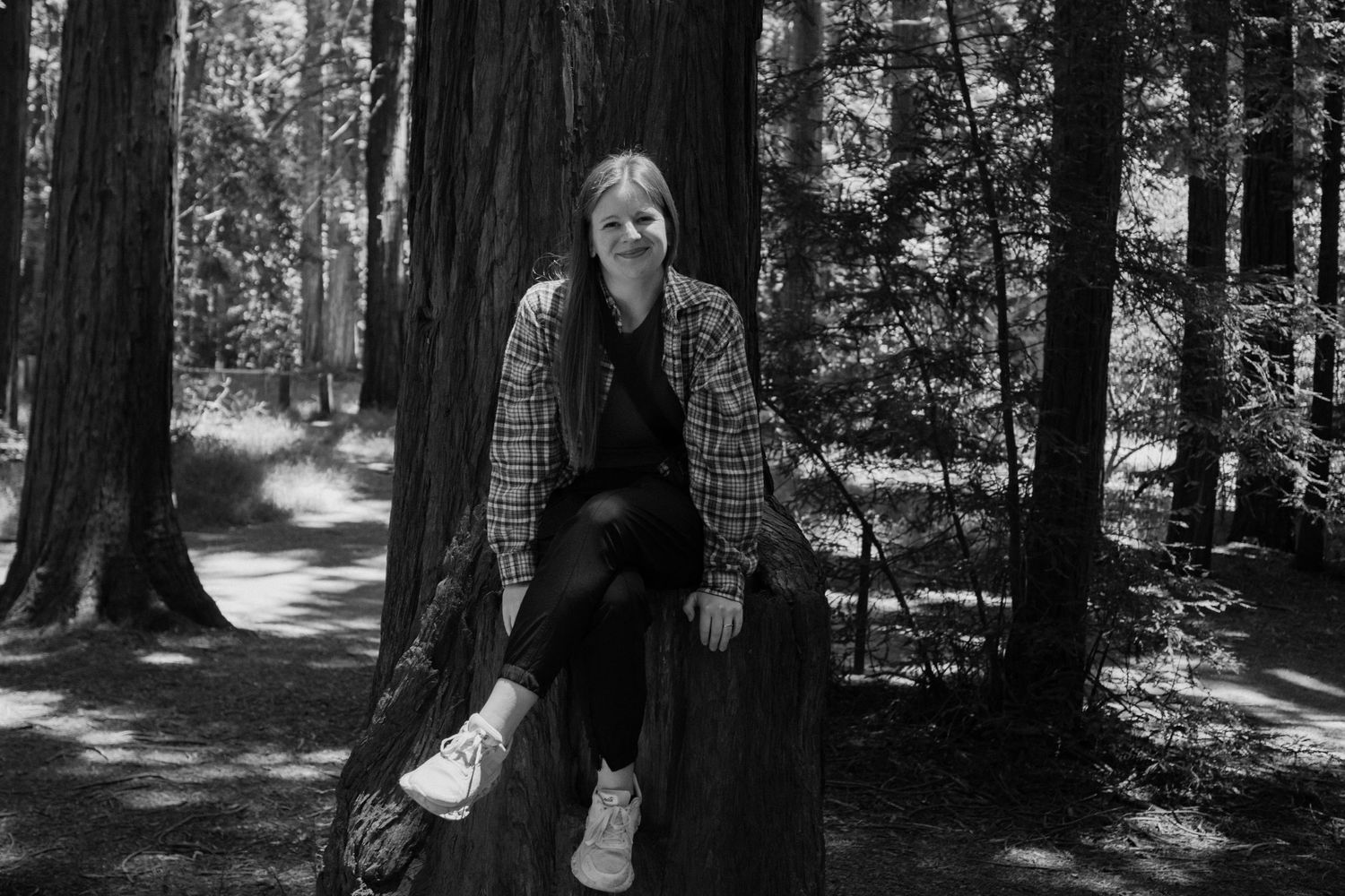 Taylor sitting on a tree stump in a forest, wearing a plaid jacket, smiling in a black and white photo.