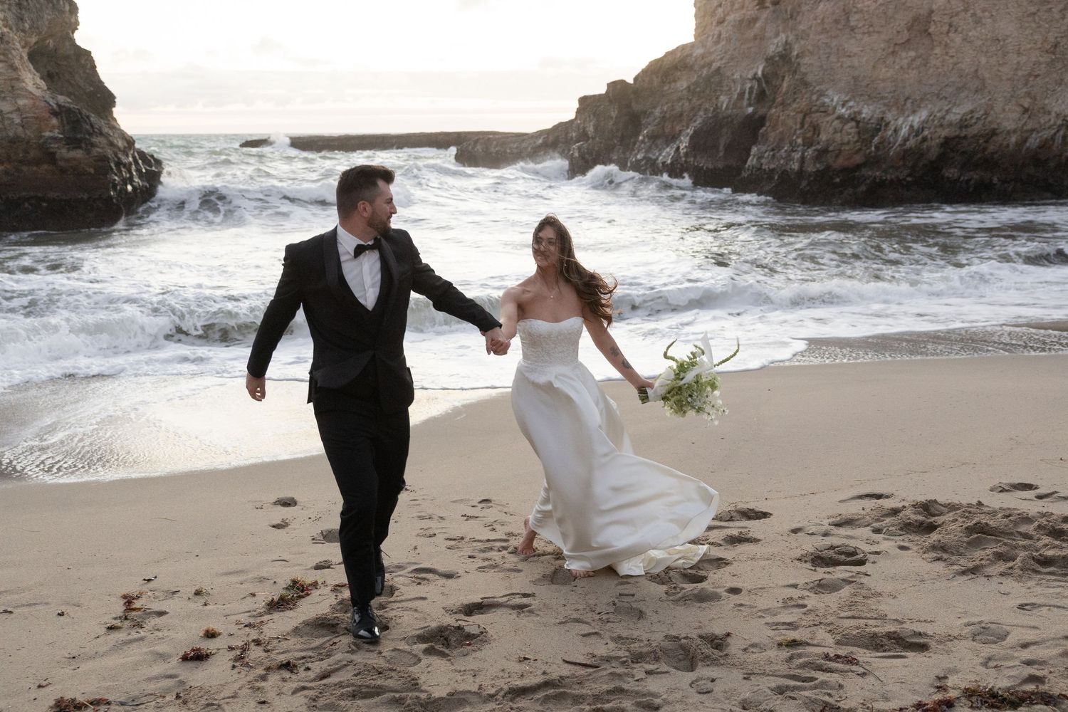 Bride and groom walk hand-in-hand on a scenic beach with crashing waves and rocky cliffs on their wedding day.