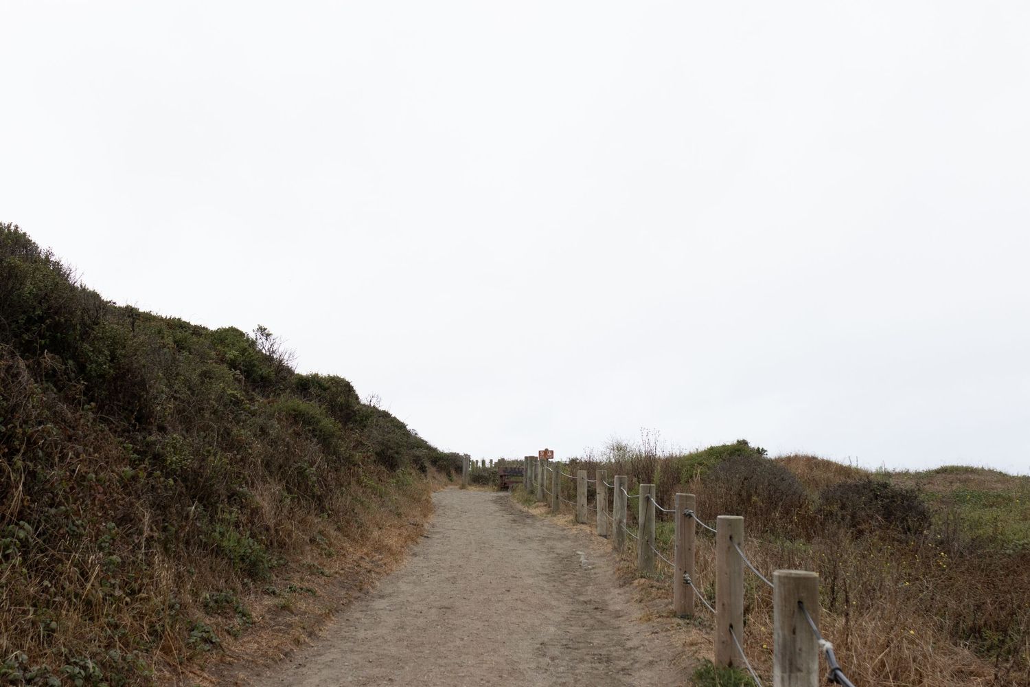 A narrow dirt path winds through coastal shrubland, flanked by wooden fence posts and overgrown hillsides under a cloudy sky.
