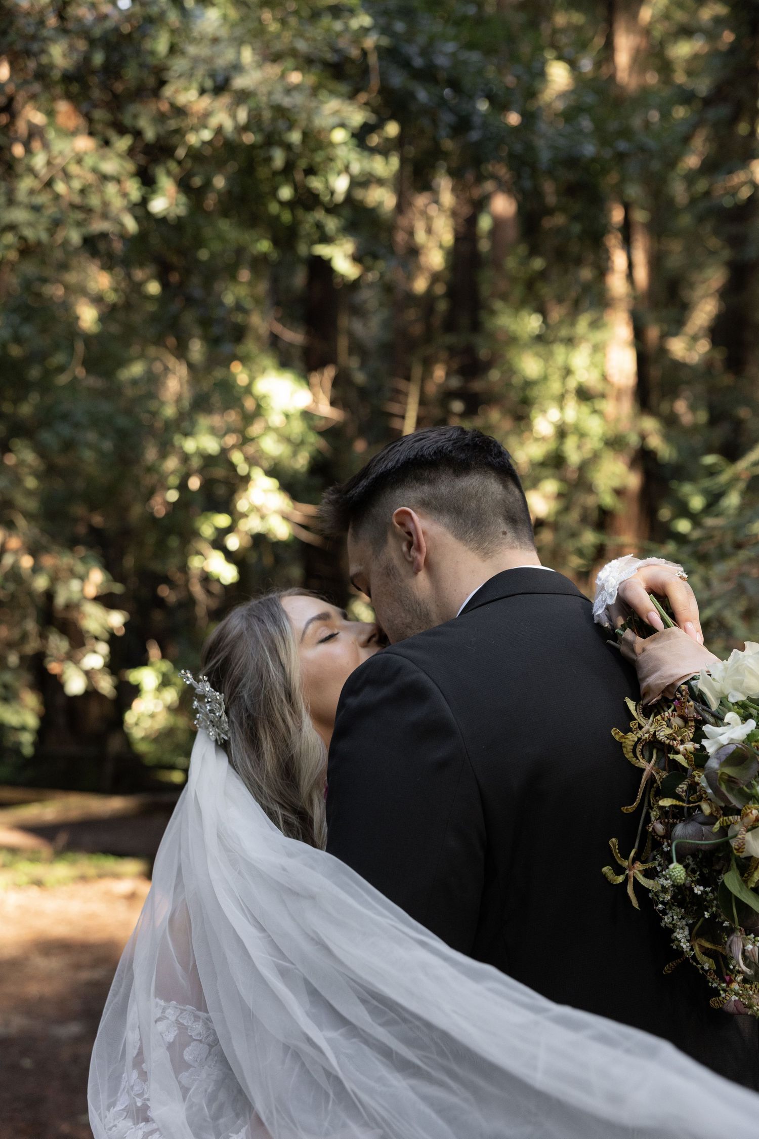 Bride and groom embrace in a sunlit redwood forest, bride holding a lush greenery bouquet by Taylor Zimmerman Photo