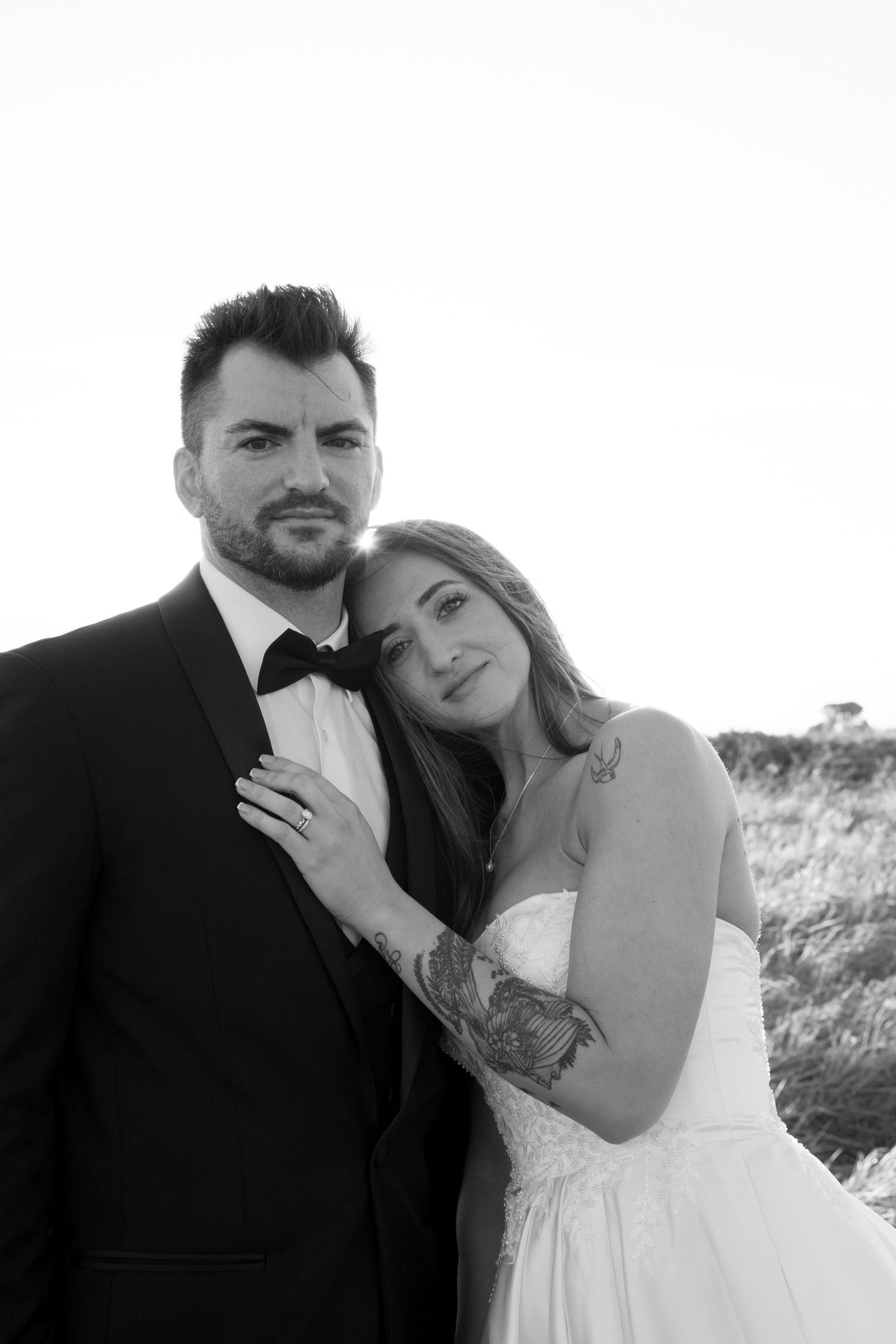 Wedding portrait of bride and groom posing outdoors, bride with tattoo in strapless gown, groom in tuxedo in black and white.
