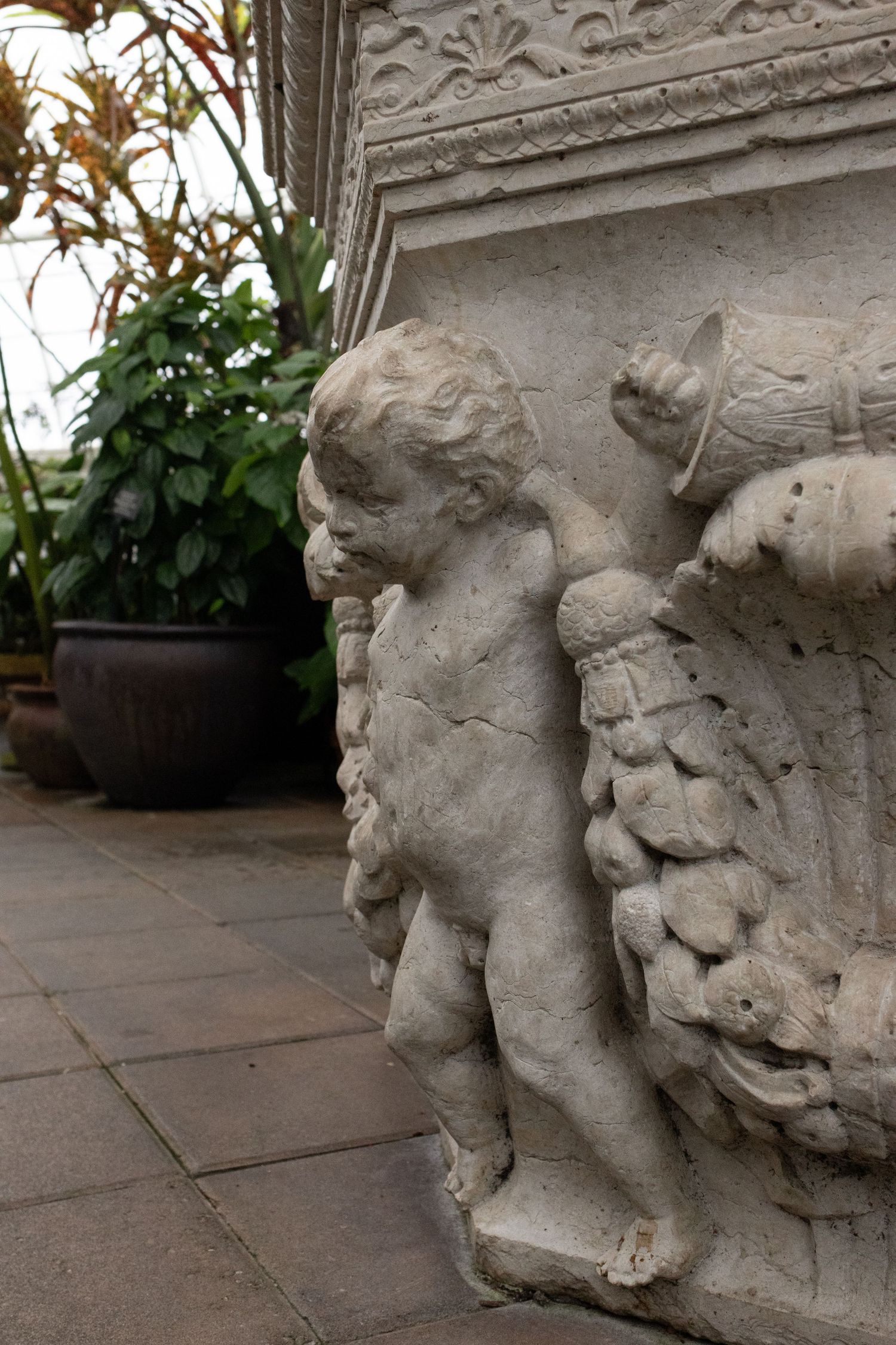 Stone cherub angel sculpture on corner of decorative pedestal with lush green potted plants in background.