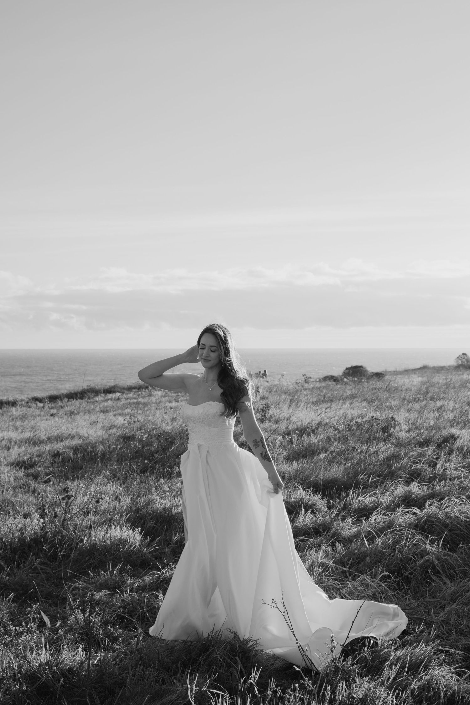 Photo of a bride in a flowing strapless gown standing on a grassy coastal hillside in black and white.