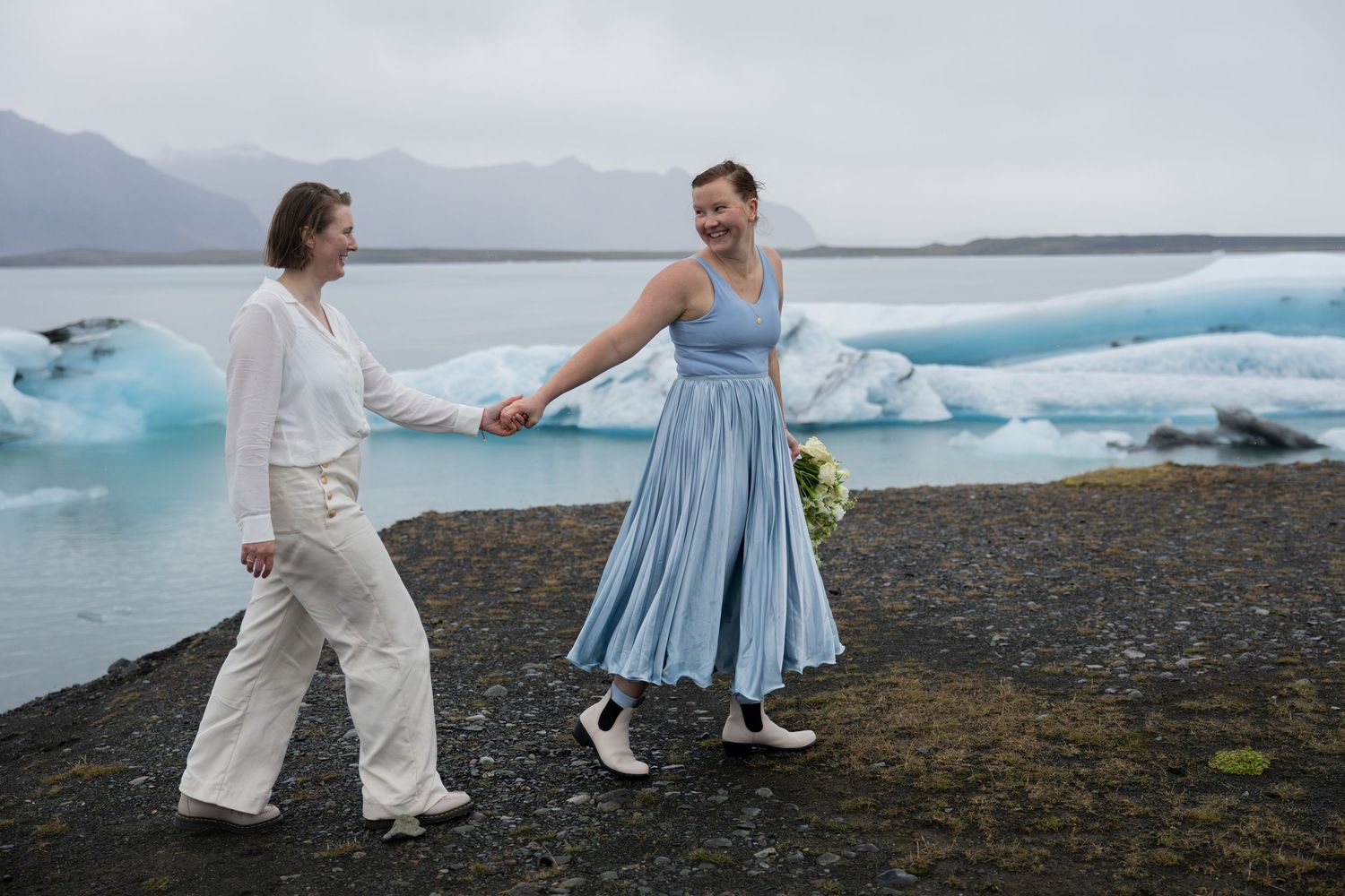 One bride leading the other along a path at Jokulsarlon glacier lagoon in Iceland with icebergs in the back by Taylor Zimmerman Photo