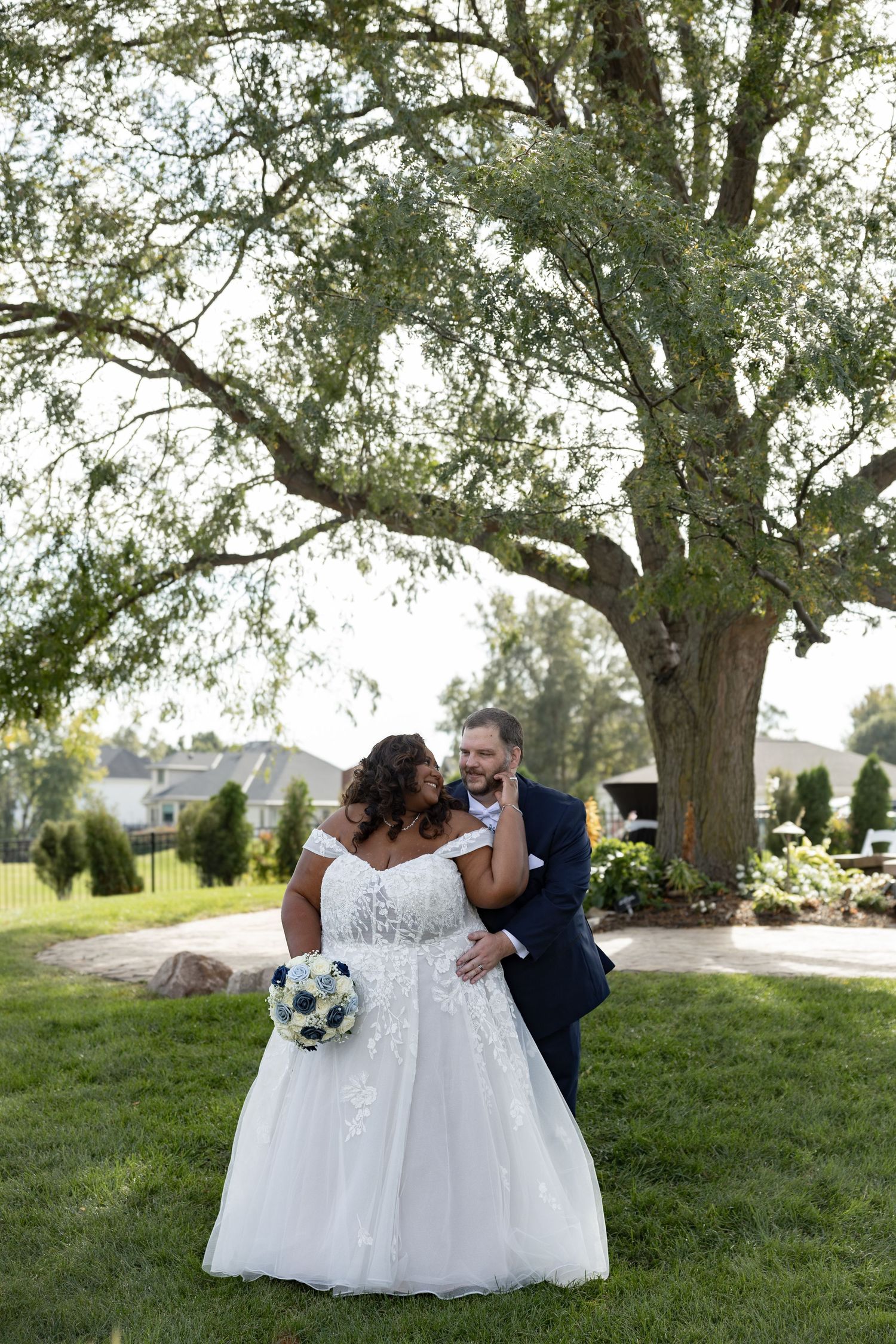 Bride in white off-shoulder gown holds bouquet as groom in navy suit embraces her under a large tree outdoors by Taylor Zimmerman Photo