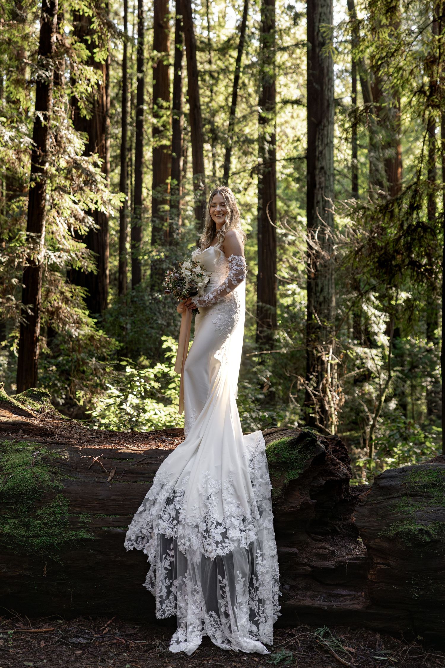 Bride in wedding gown holding bouquet, standing on fallen log in lush redwood forest by Taylor Zimmerman Photo