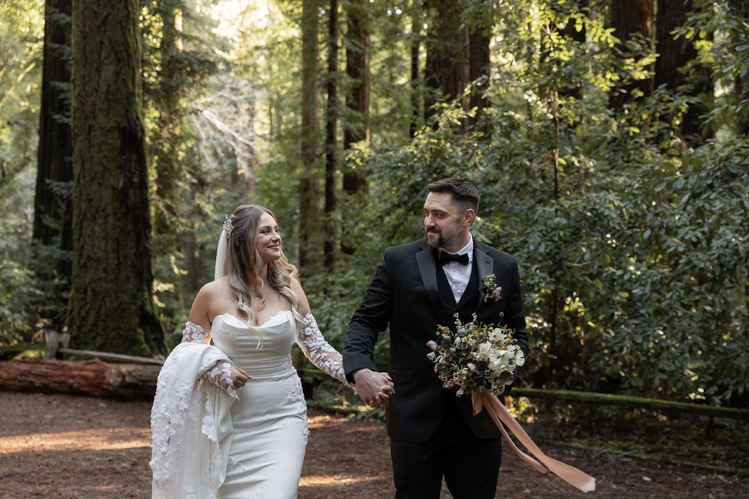 Bride and groom walk hand-in-hand through a lush redwood forest, smiling joyfully on their wedding day by Taylor Zimmerman Photo