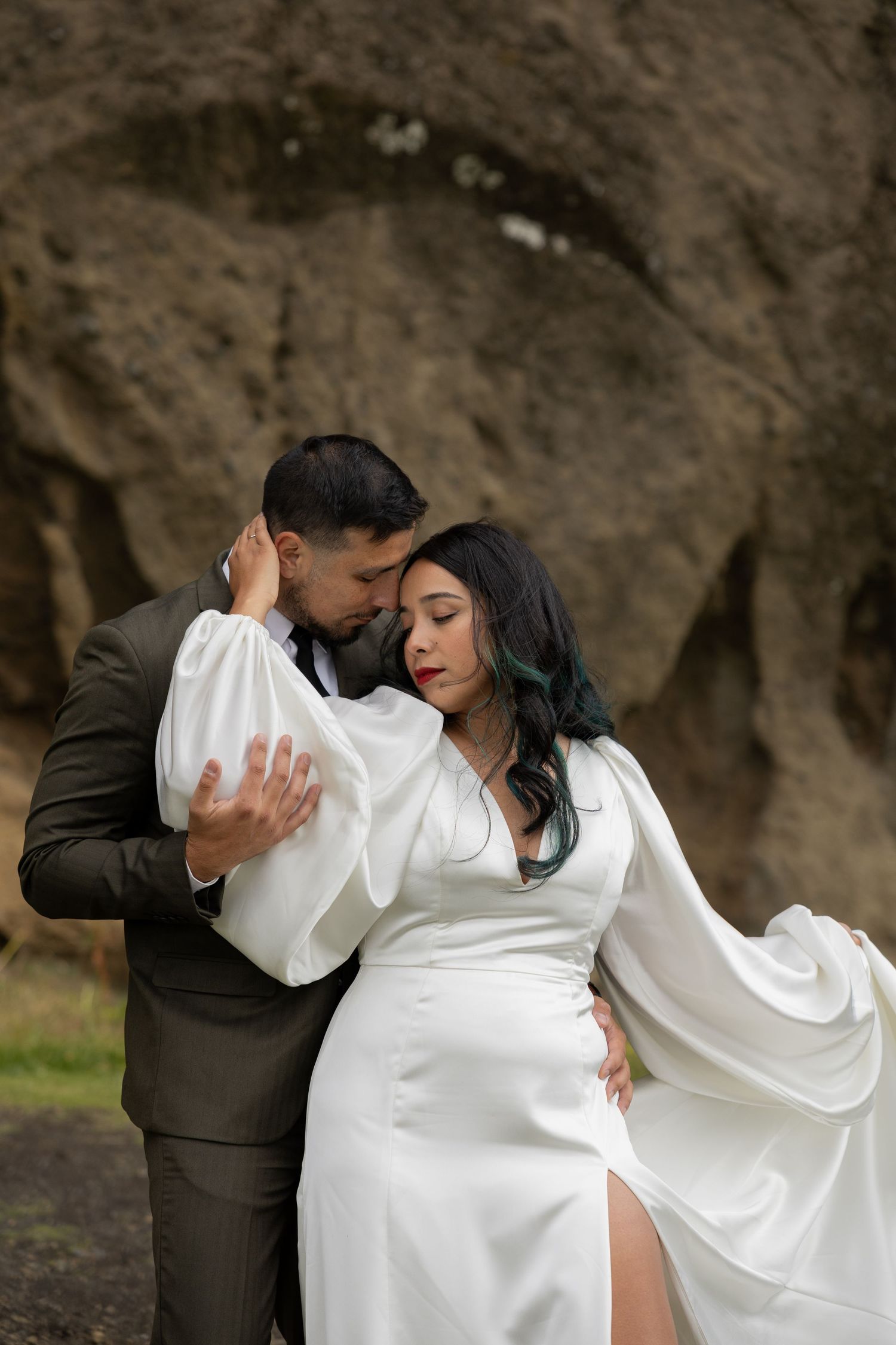 Bride in white satin gown and groom in olive suit pose before rocky outdoor backdrop in Iceland by Taylor Zimmerman Photo