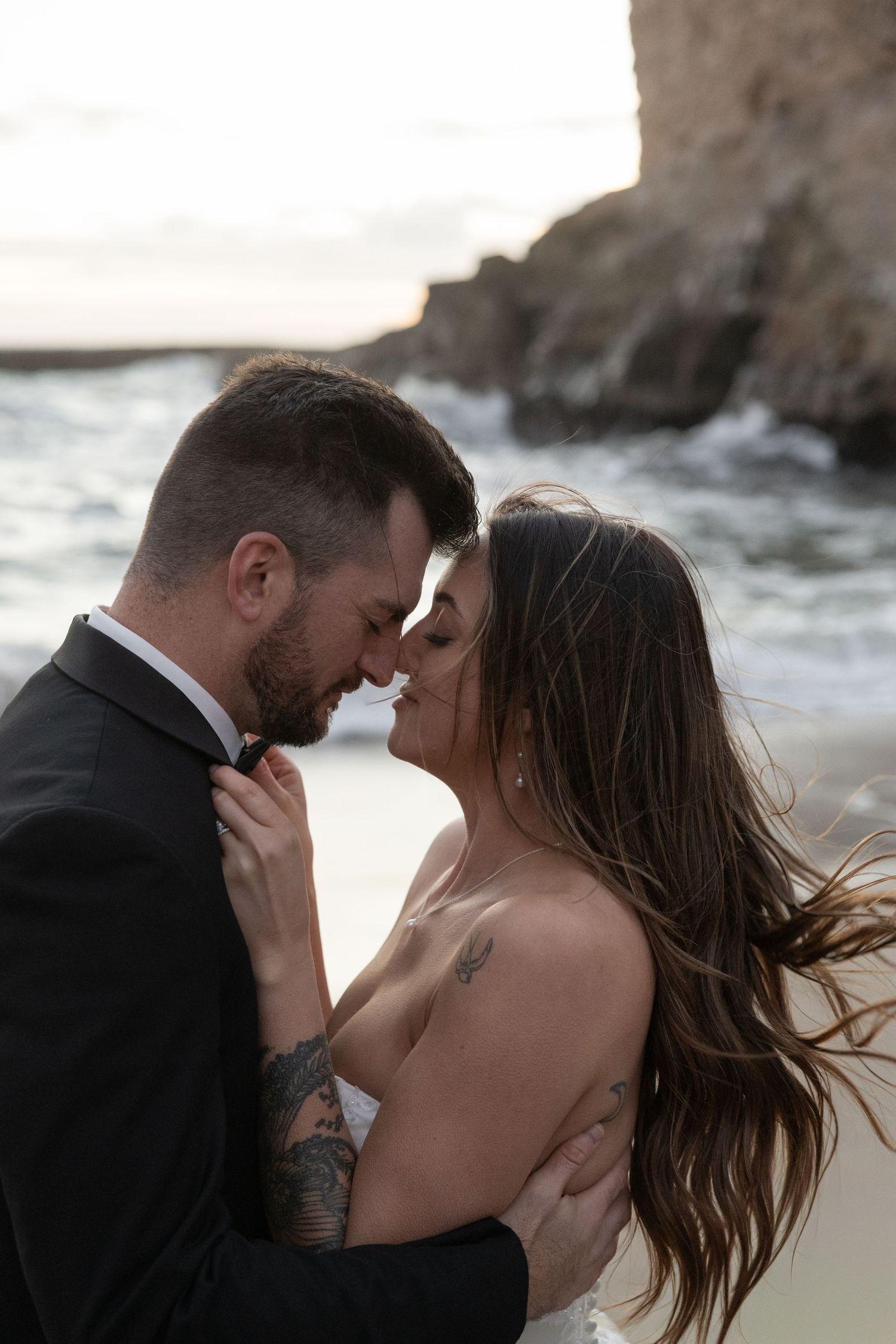 A bride and groom share a romantic beach side moment, foreheads touching, dressed in wedding attire with ocean waves behind them.
