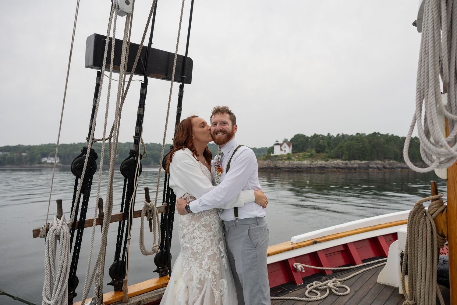 Bride kisses her husband on the cheek on a sailboat deck with the ocean and a lighthouse in the background by Taylor Zimmerman Photo