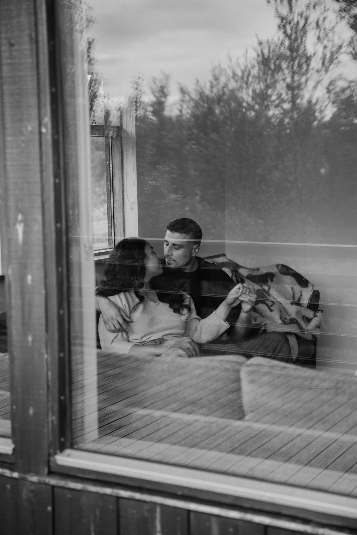 A couple holding hands on a couch, viewed through a window with trees reflected outside in black and white by Taylor Zimmerman Photo