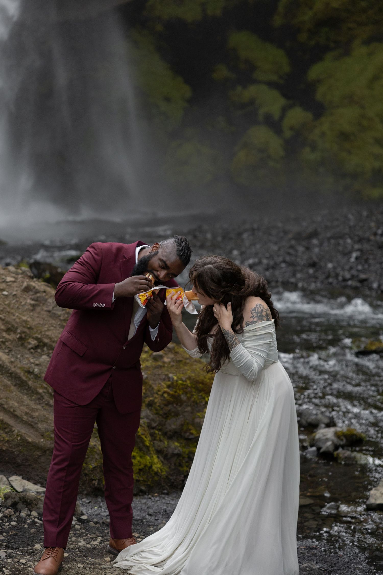 A bride in a flowy dress and groom in a maroon suit eat hot dogs in front of an Iceland waterfall on their elopement day by Taylor Zimmerman