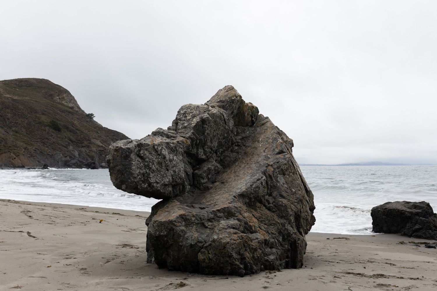 Large weathered rock formation on a sandy beach with ocean waves and misty coastal cliffs in the background.