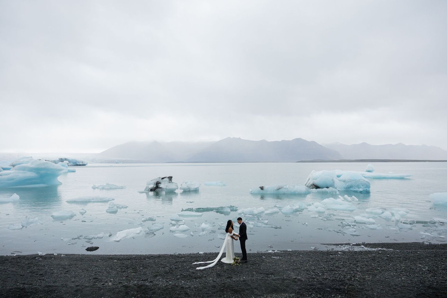Couple in wedding attire standing on black sand beach at Jokulsarlon glacier lagoon in Iceland with icebergs by Taylor Zimmerman Photo
