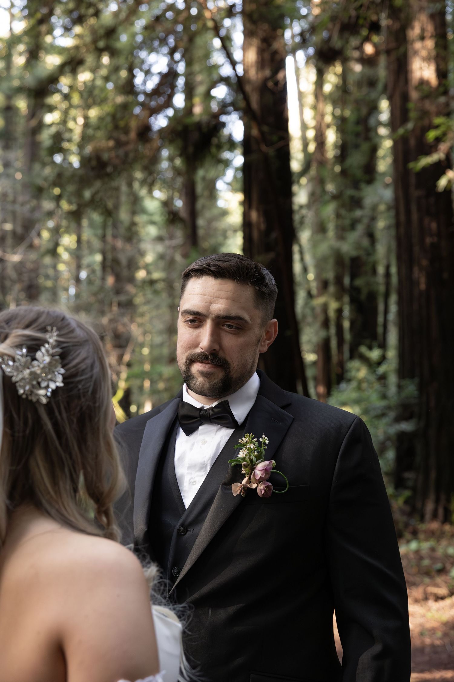 Groom in black tuxedo with floral boutonniere stands facing bride during redwood wedding ceremony by Taylor Zimmerman Photo