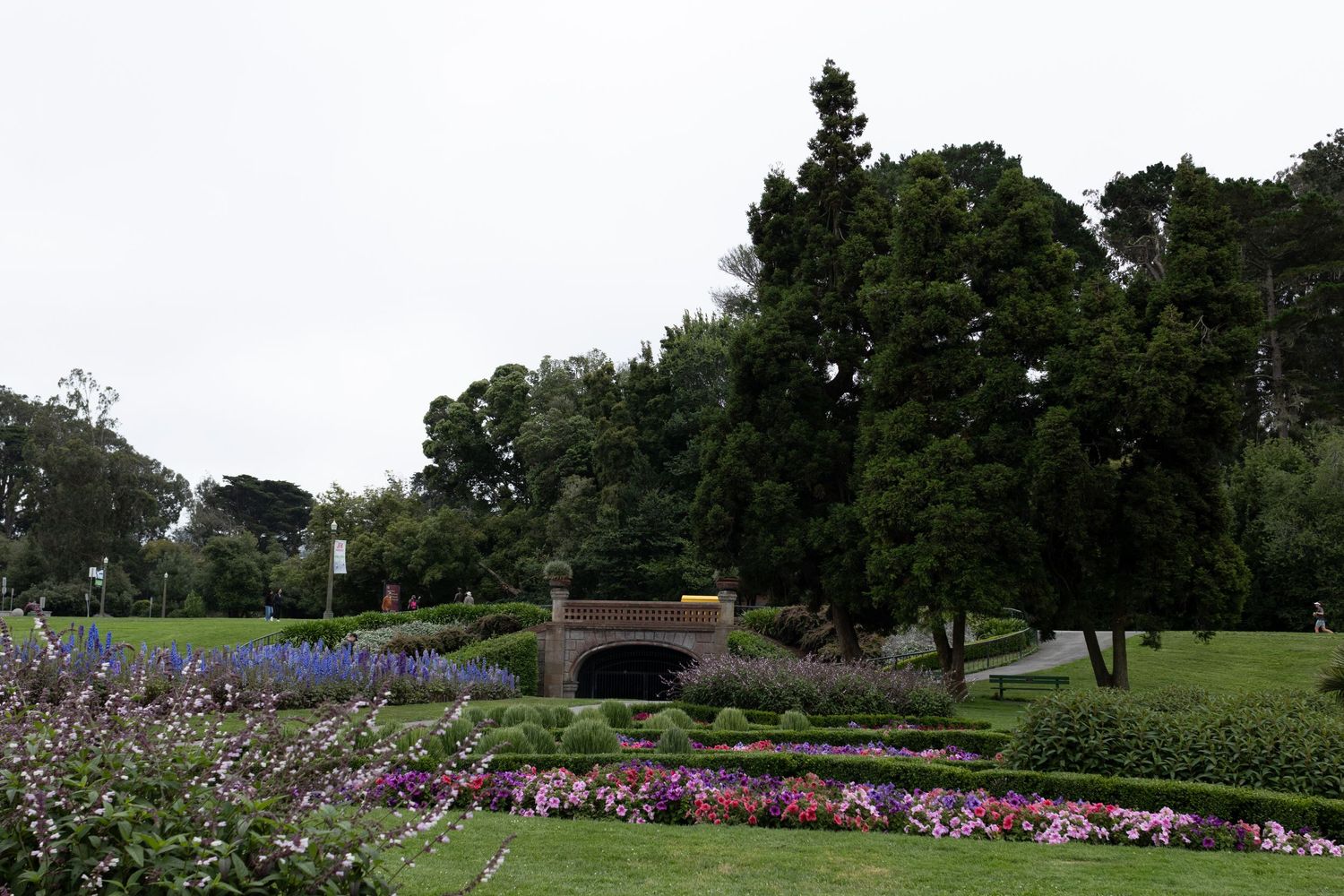 Lush park garden with colorful flower beds, brick archway, and tall green trees along the JFK promenade.