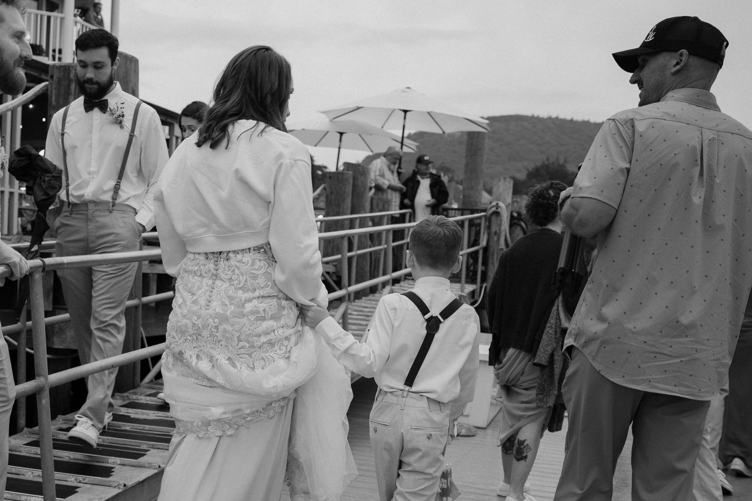 Bride holding hands with young boy in suspenders walking to a sailboat in black and white by Taylor Zimmerman Photo