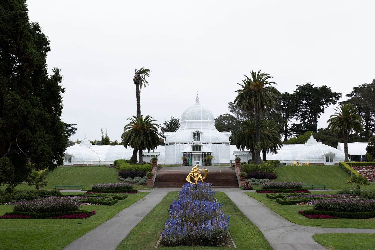 The Conservatory of Flowers surrounded by palm trees, colorful flower gardens, and manicured hedges on an overcast day.