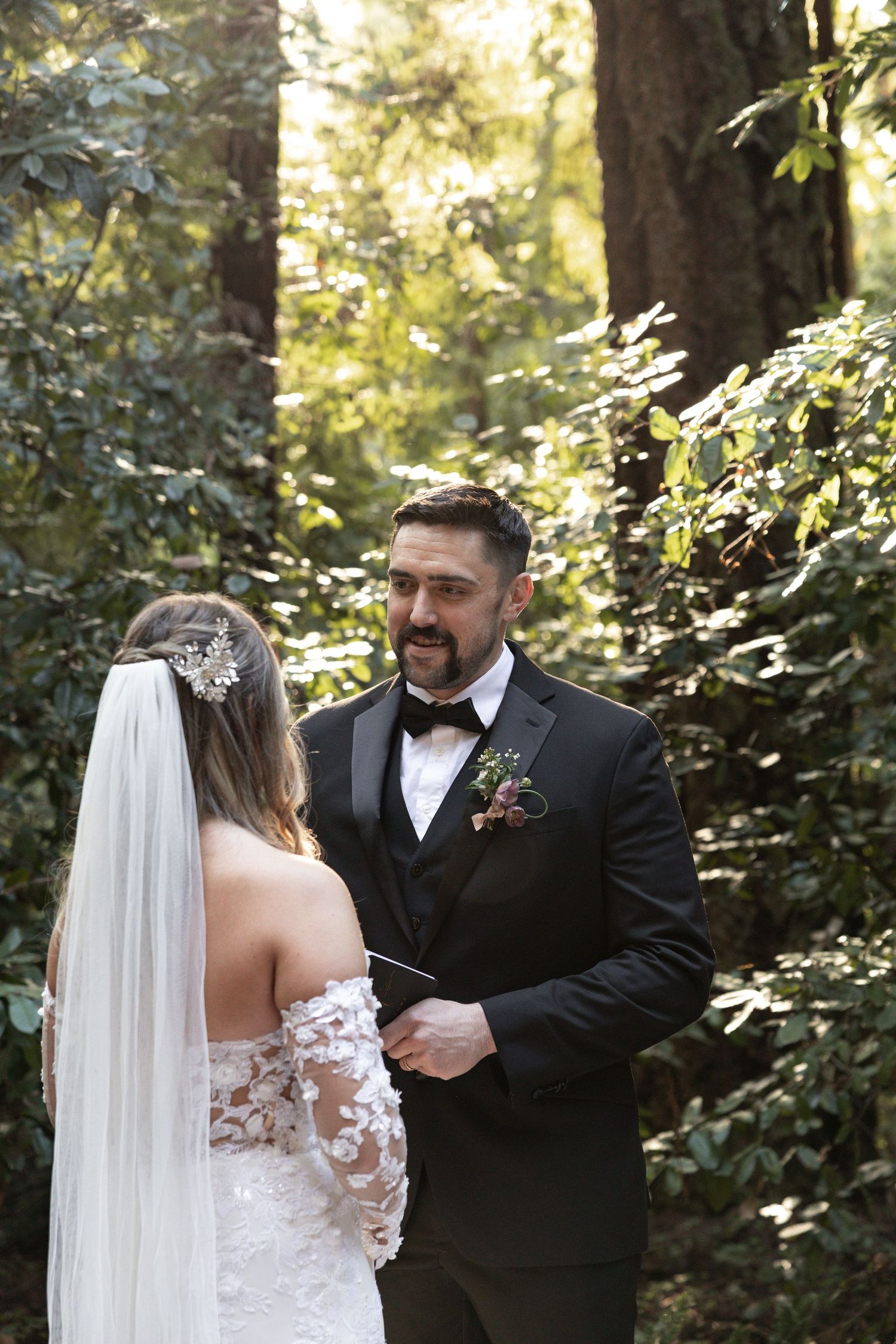 Groom in black tuxedo reads vows to bride in lace off-shoulder gown during outdoor redwood wedding ceremony by Taylor Zimmerman Photo