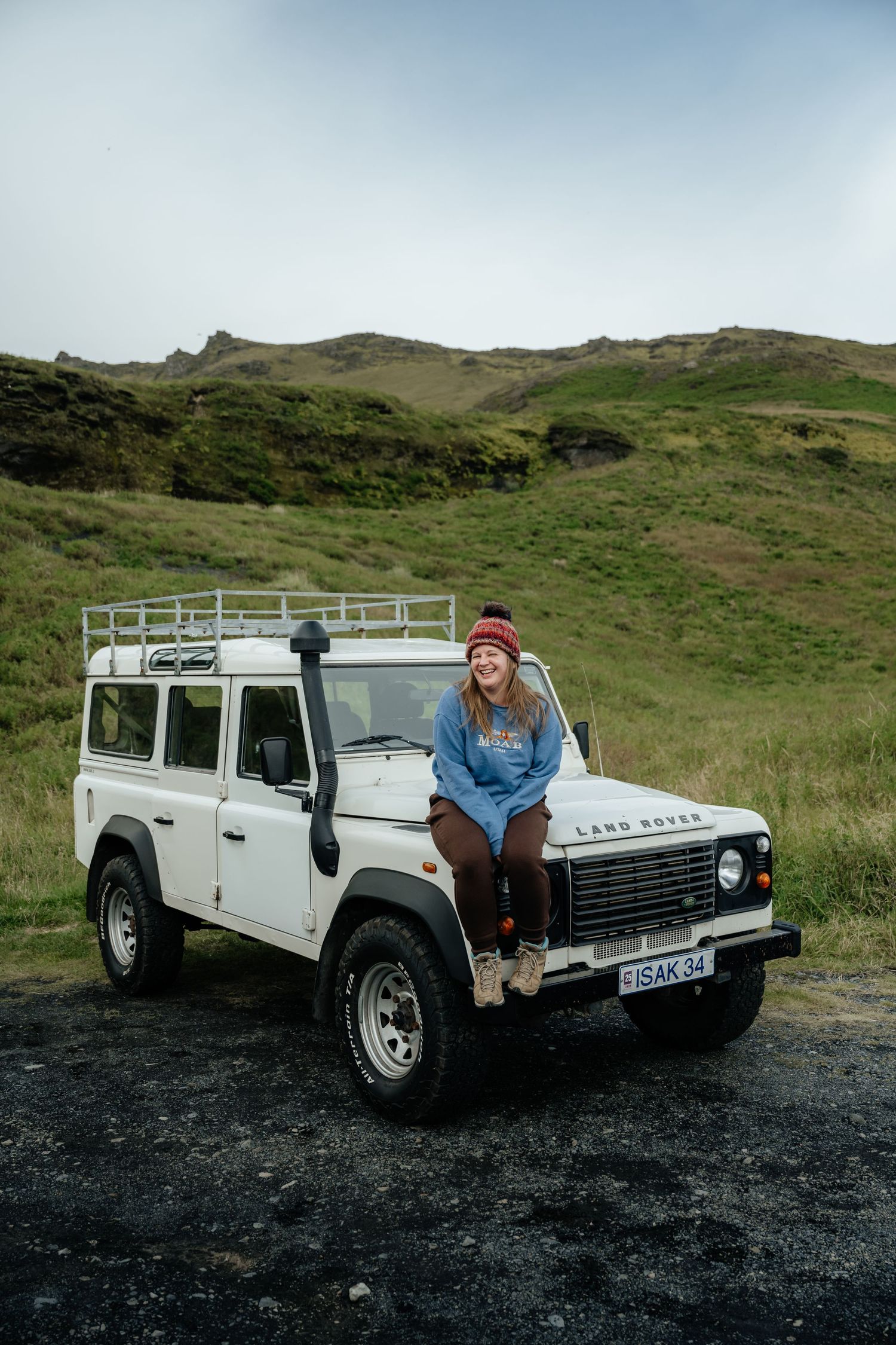Woman in red beanie sitting on white Land Rover Defender hood with green hills in background.