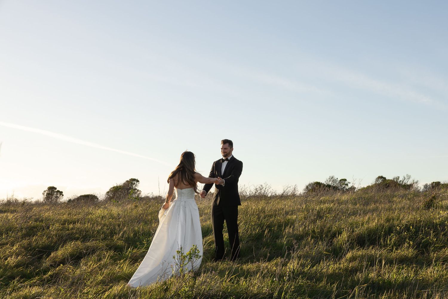 A bride and groom hold hands in a sunlit green field during their outdoor wedding ceremony.