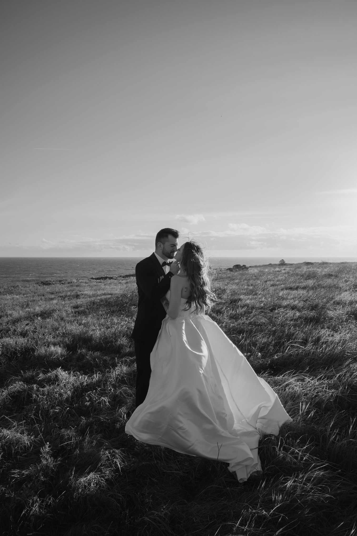 Bride and groom embracing in a windswept field by the ocean on their wedding day in black and white.
