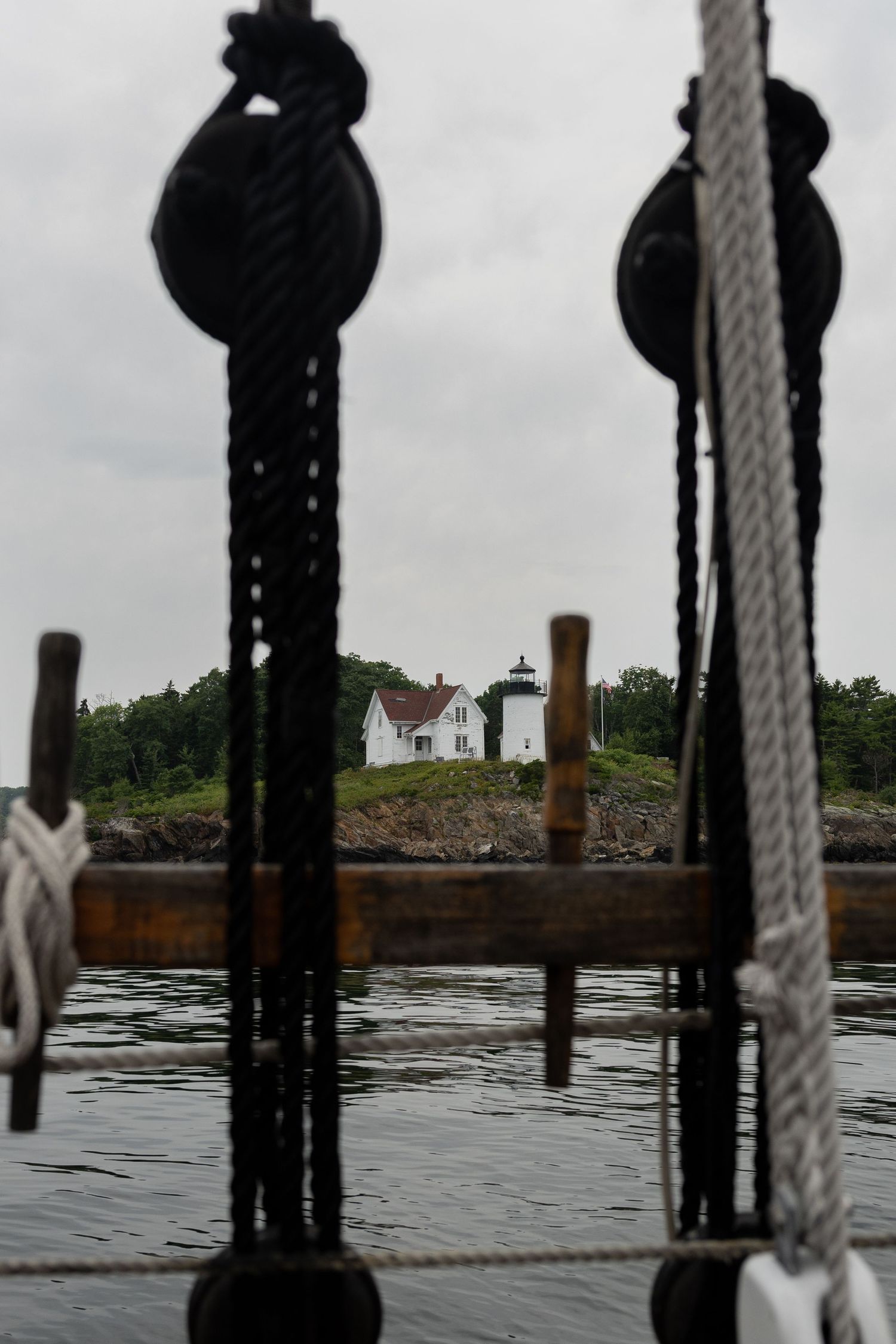 A lighthouse viewed through tall ship rigging and pulleys on a rocky Maine coastline under overcast skies by Taylor Zimmerman Photo