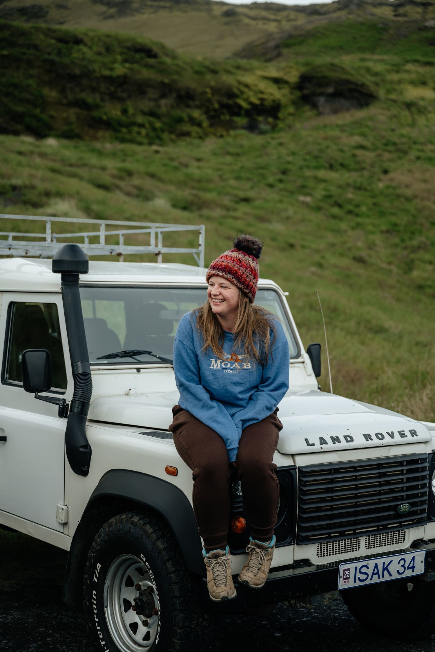 Woman in blue sweatshirt and red beanie sitting on white Land Rover Defender hood in green Icelandic landscape