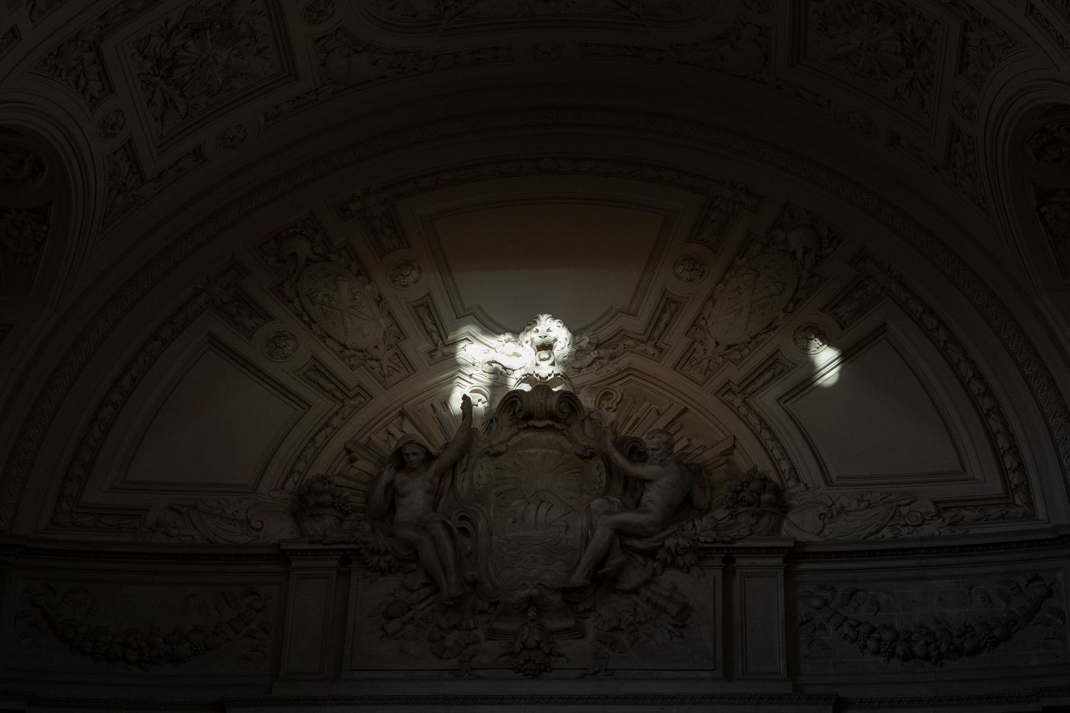 Ornate baroque ceiling with sculpted cherubs and coat of arms in dimly lit historic building interior.