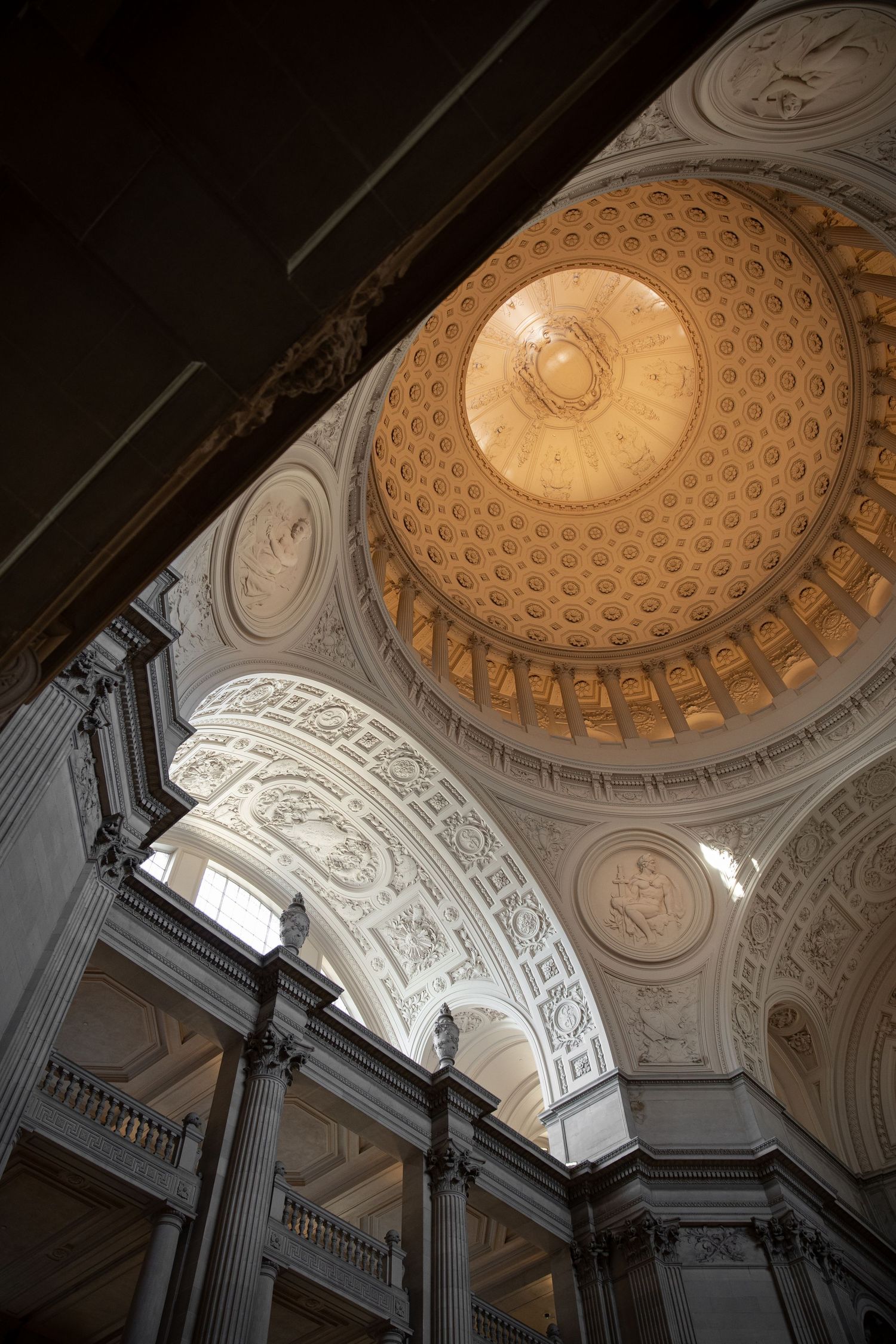 Interior view of a grand neoclassical dome with ornate coffered ceiling, decorative arches, and tall columns in warm golden light.