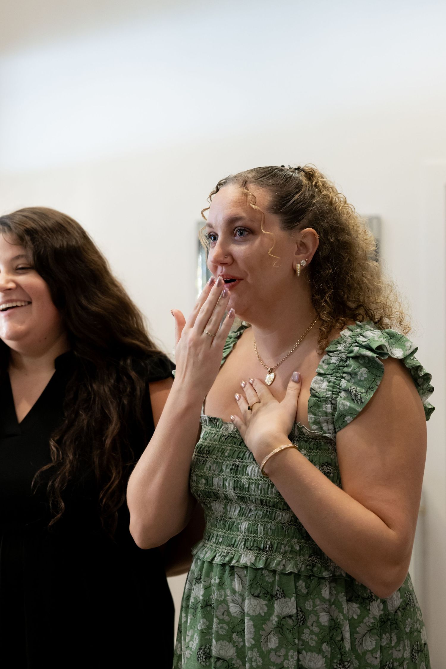 Two women smiling, one in green floral dress with hand over mouth in surprise, another in black dress behind her by Taylor Zimmerman Photo