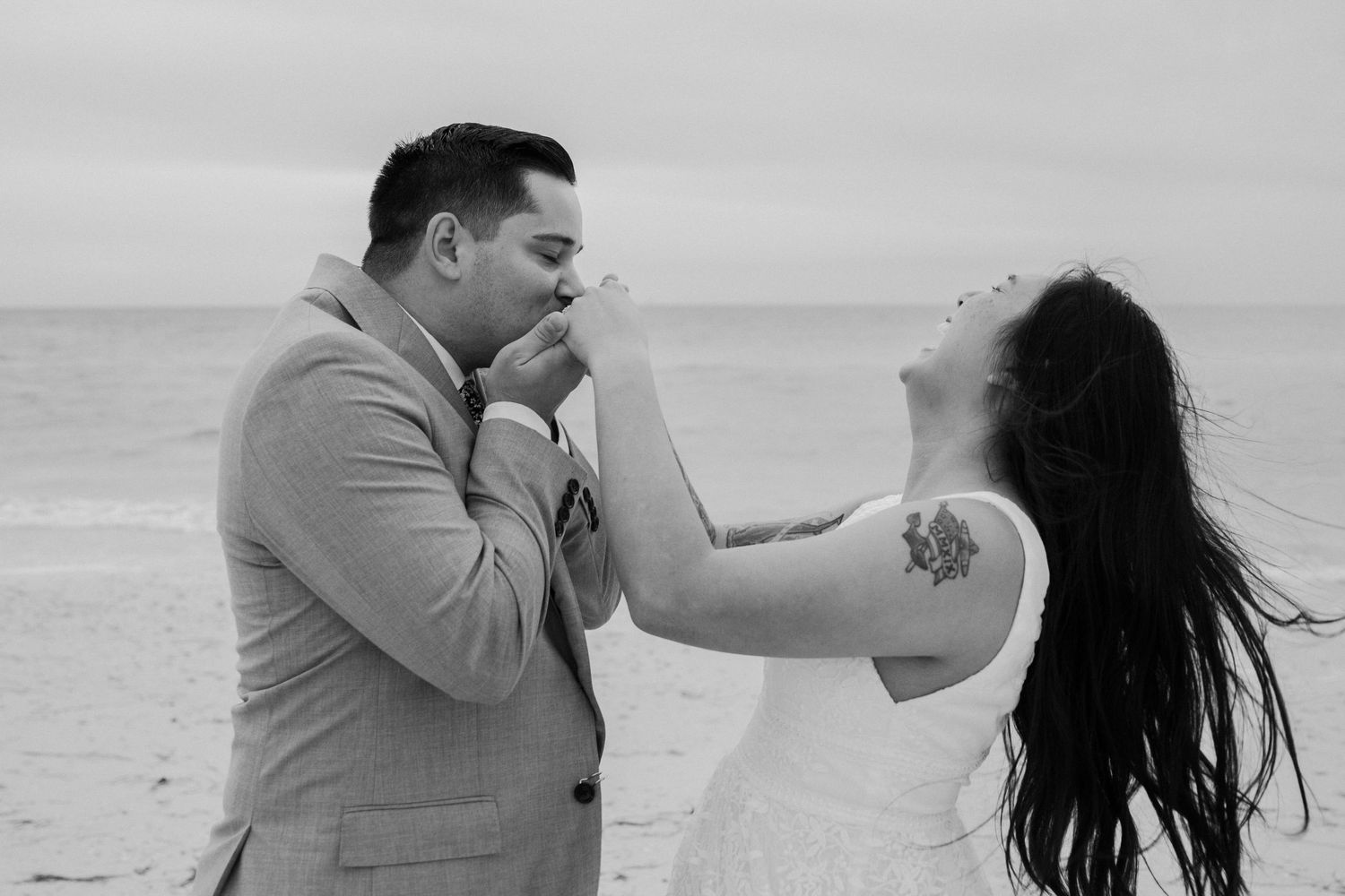 Groom kissing bride's hands while she trows her head back laughing on a Florida beach in black and white by Taylor Zimmerman Photo